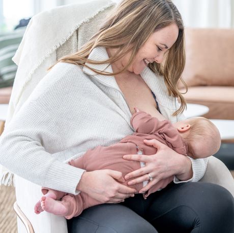 Woman in a white sweater smiling while breastfeeding a baby in a pink outfit on a chair