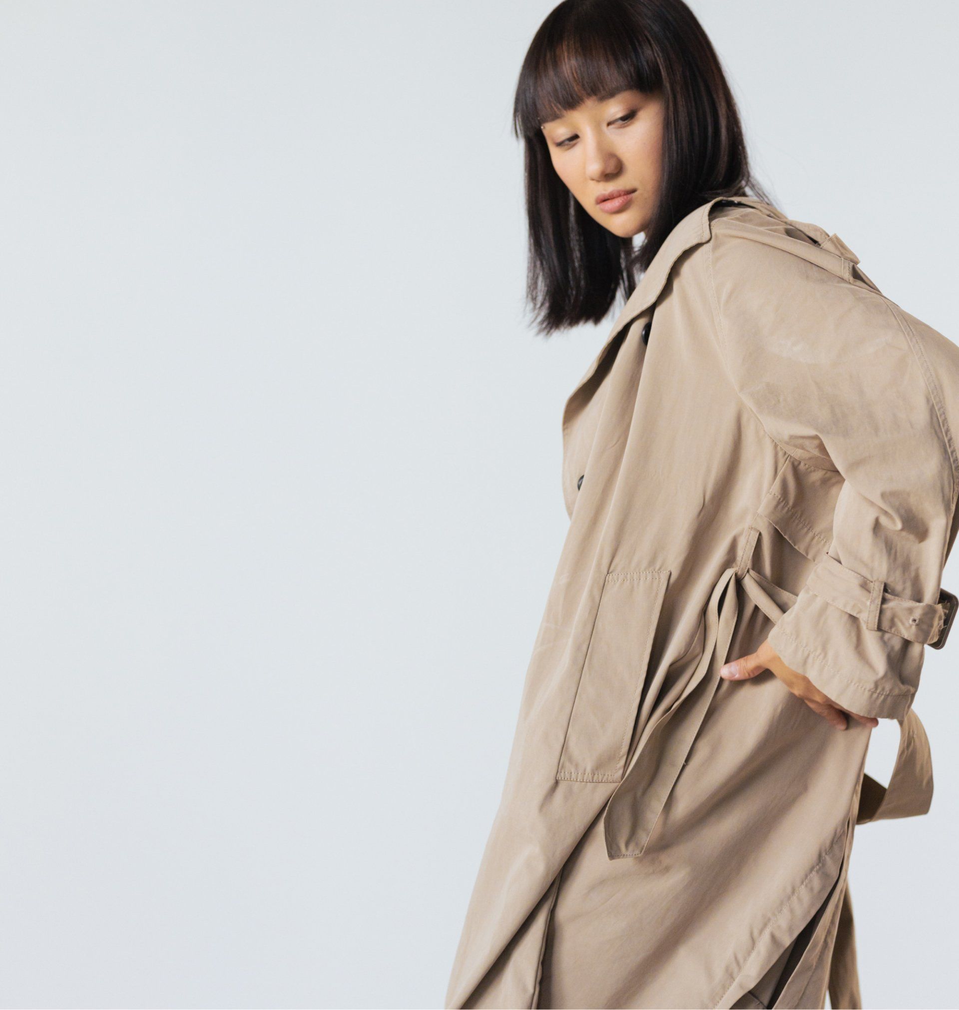 Woman wearing a tan trench coat, looking down. One hand in pocket, against a plain background.