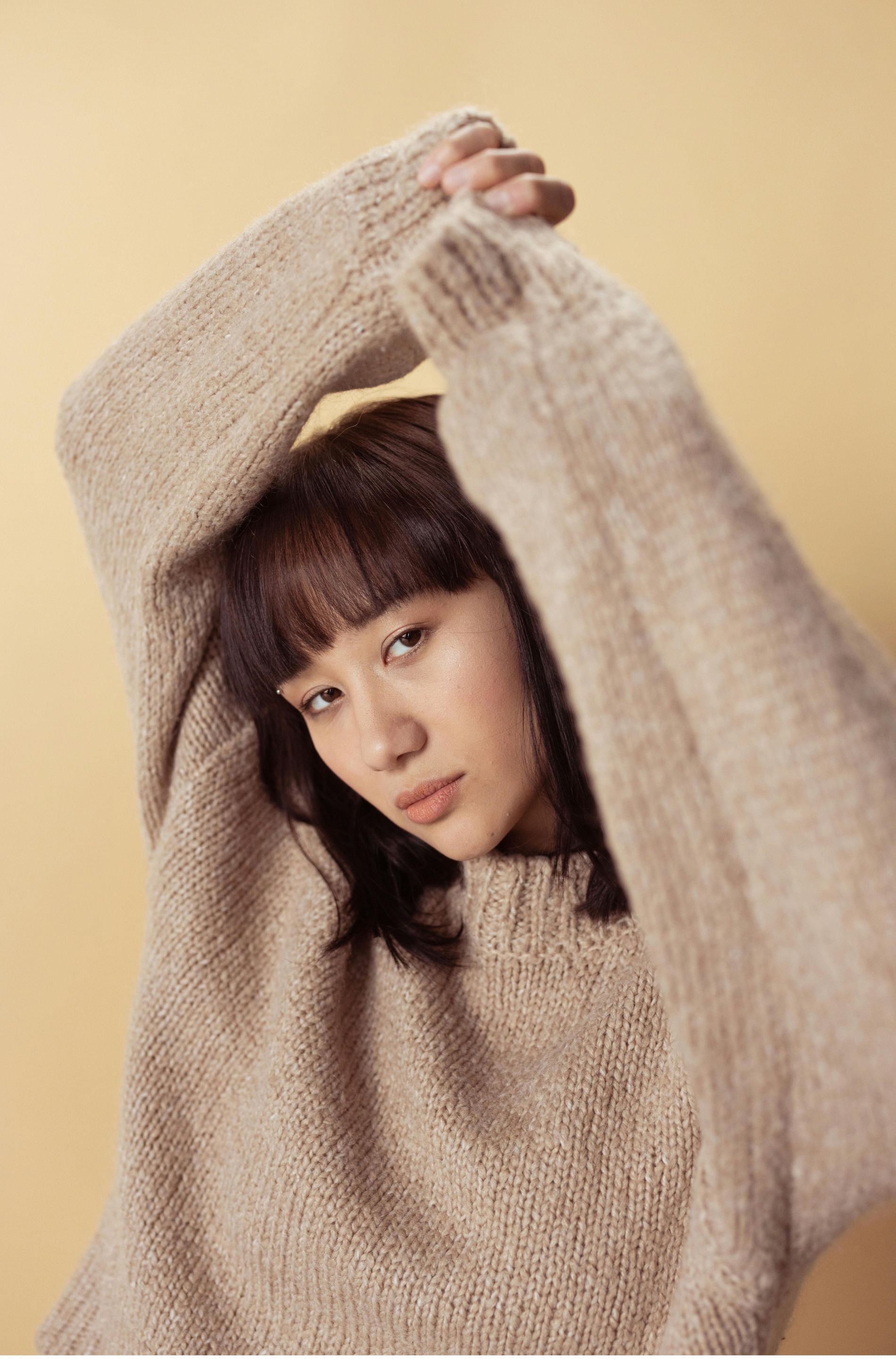 Woman in beige sweater, arms raised, looking at the camera against a light yellow background.