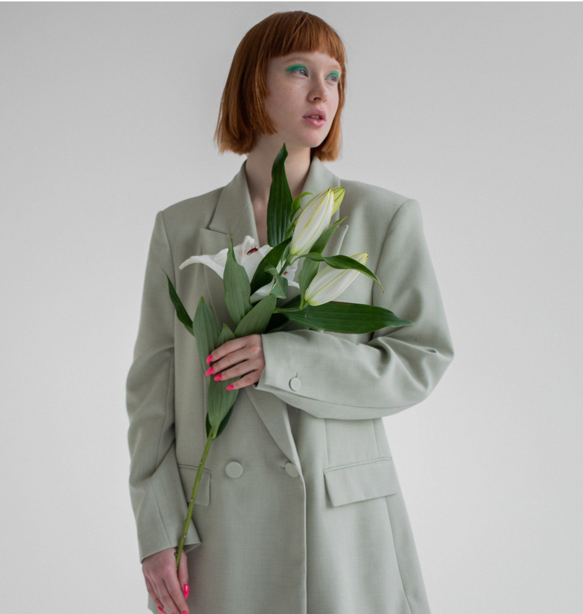 Woman in sage green blazer holds white lilies against a white backdrop.