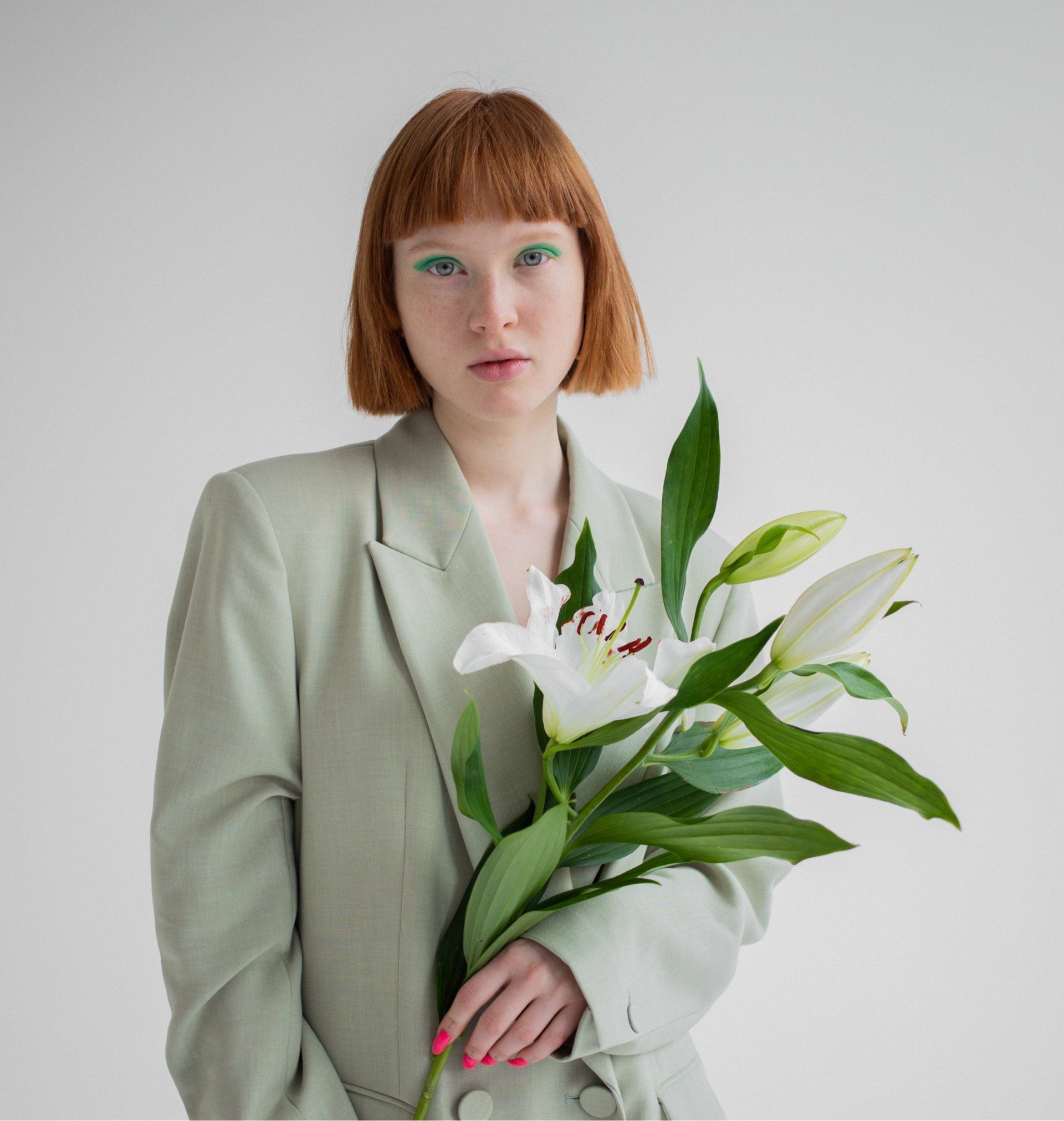 Woman in sage green blazer holds white lilies against a white backdrop.