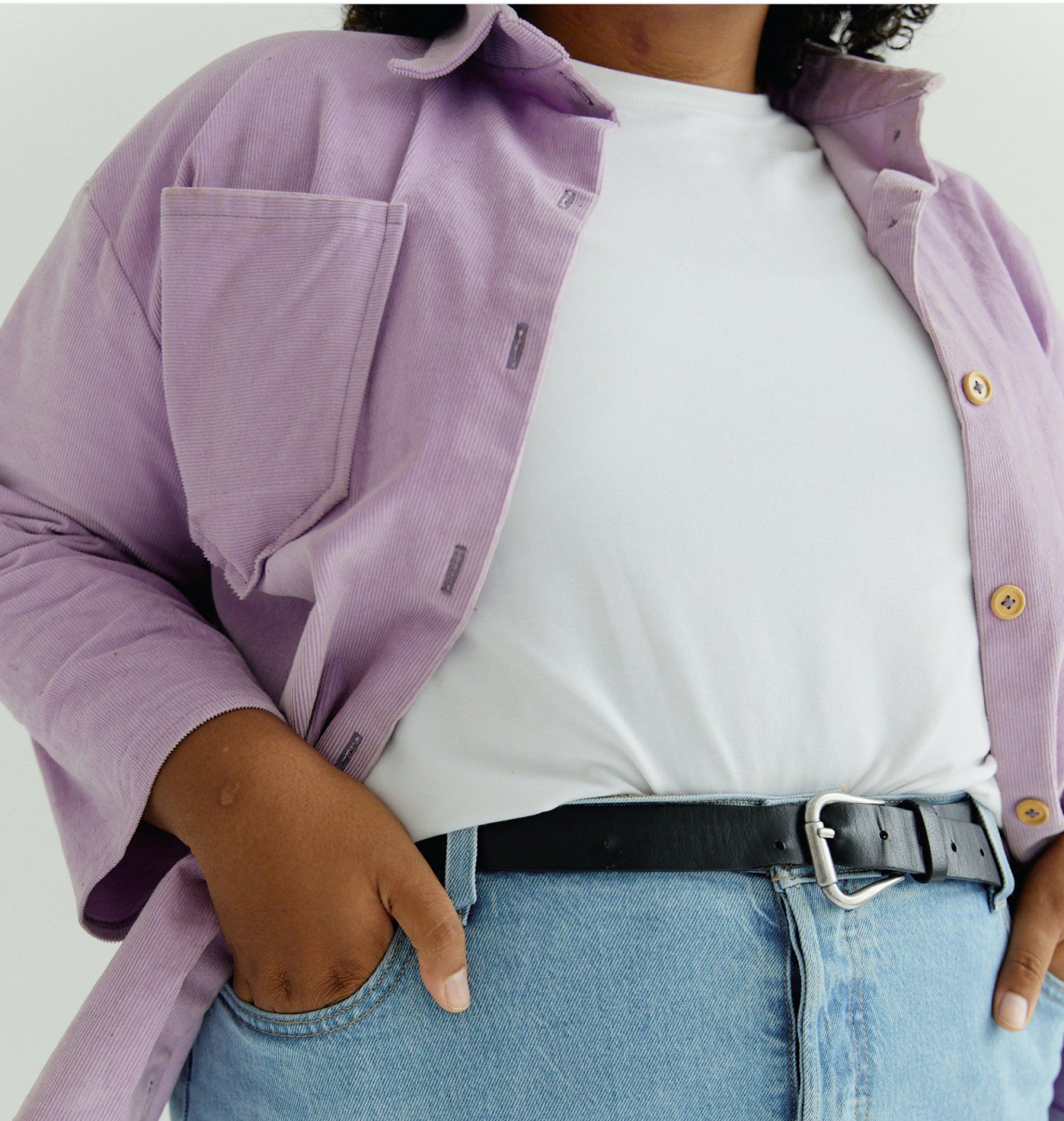 Person in a lilac shirt, white tee, and jeans looking to the side. Hands near belt, neutral expression.