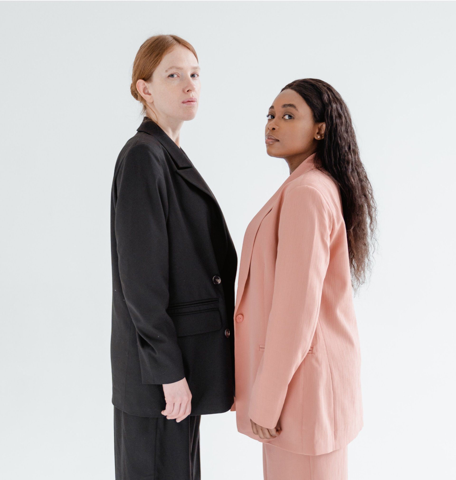 Two people in suits stand shoulder-to-shoulder, looking at the camera. One in black, the other in pink. White background.