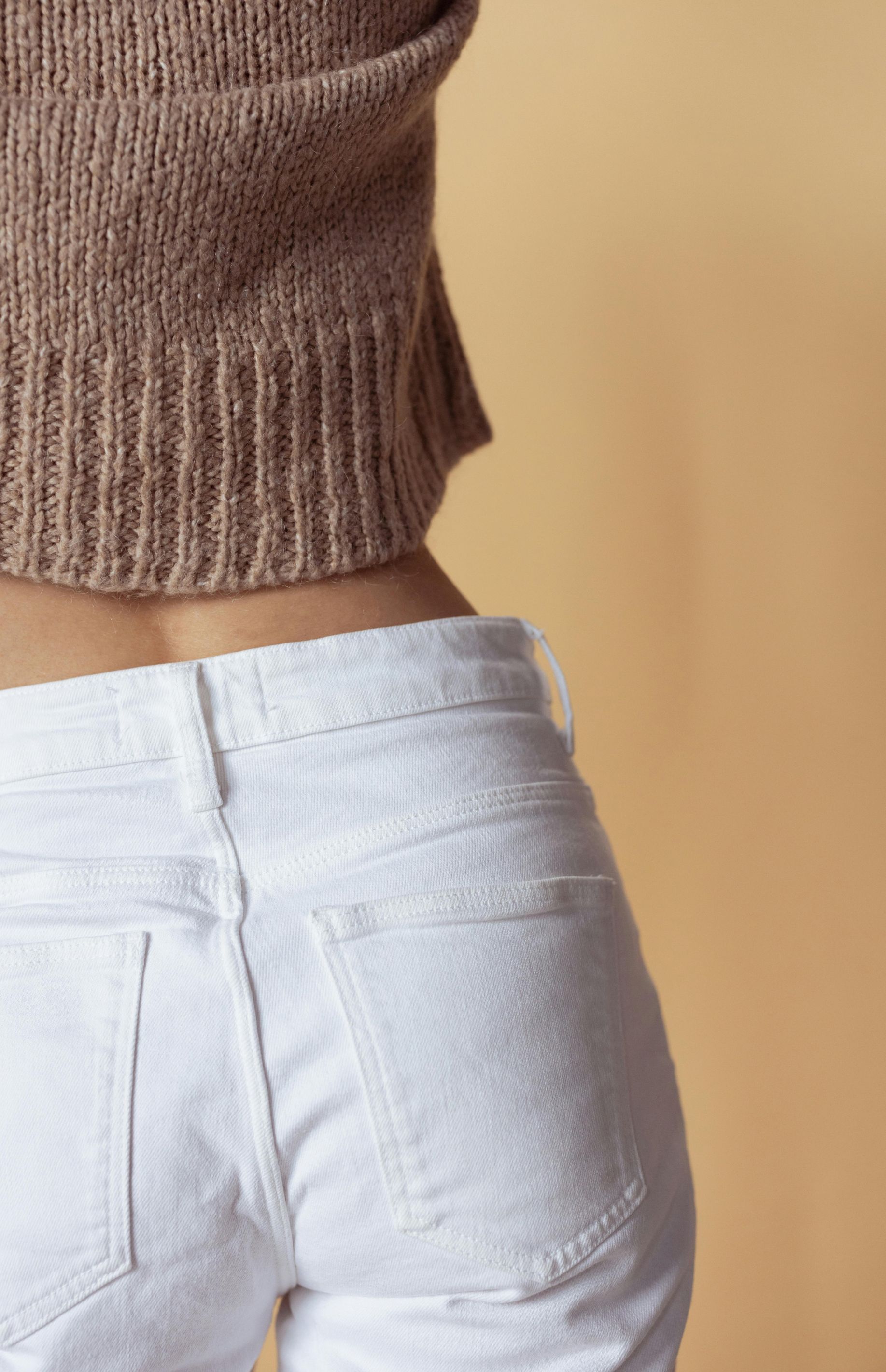 Person wearing a brown sweater and white jeans, standing against a beige background.