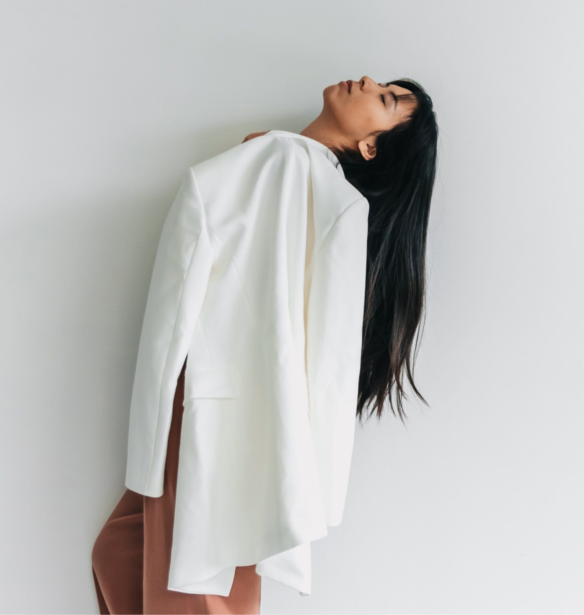 Woman wearing a white jacket, tilting head back. Long black hair, neutral expression, and pink pants. Leaning against a white wall.