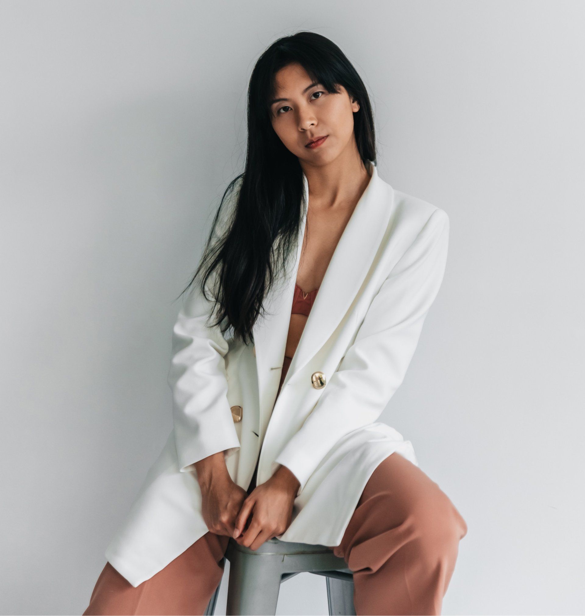 Woman wearing a white jacket, tilting head back. Long black hair, neutral expression, and pink pants. Leaning against a white wall.