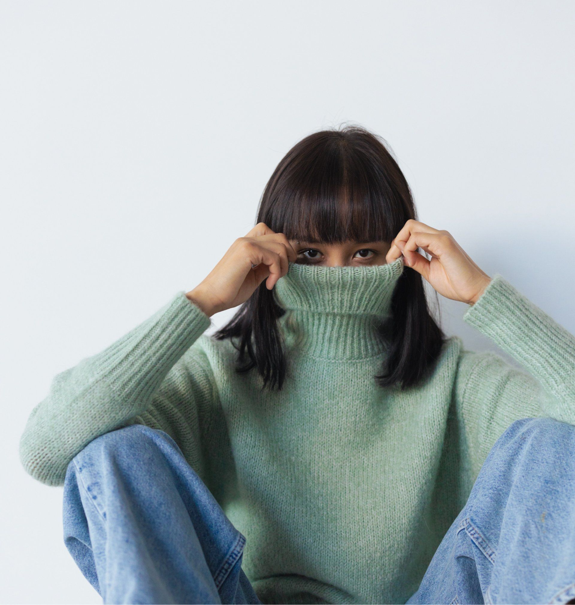 Person in green turtleneck pulls it up, partially covering face; seated, wearing jeans.