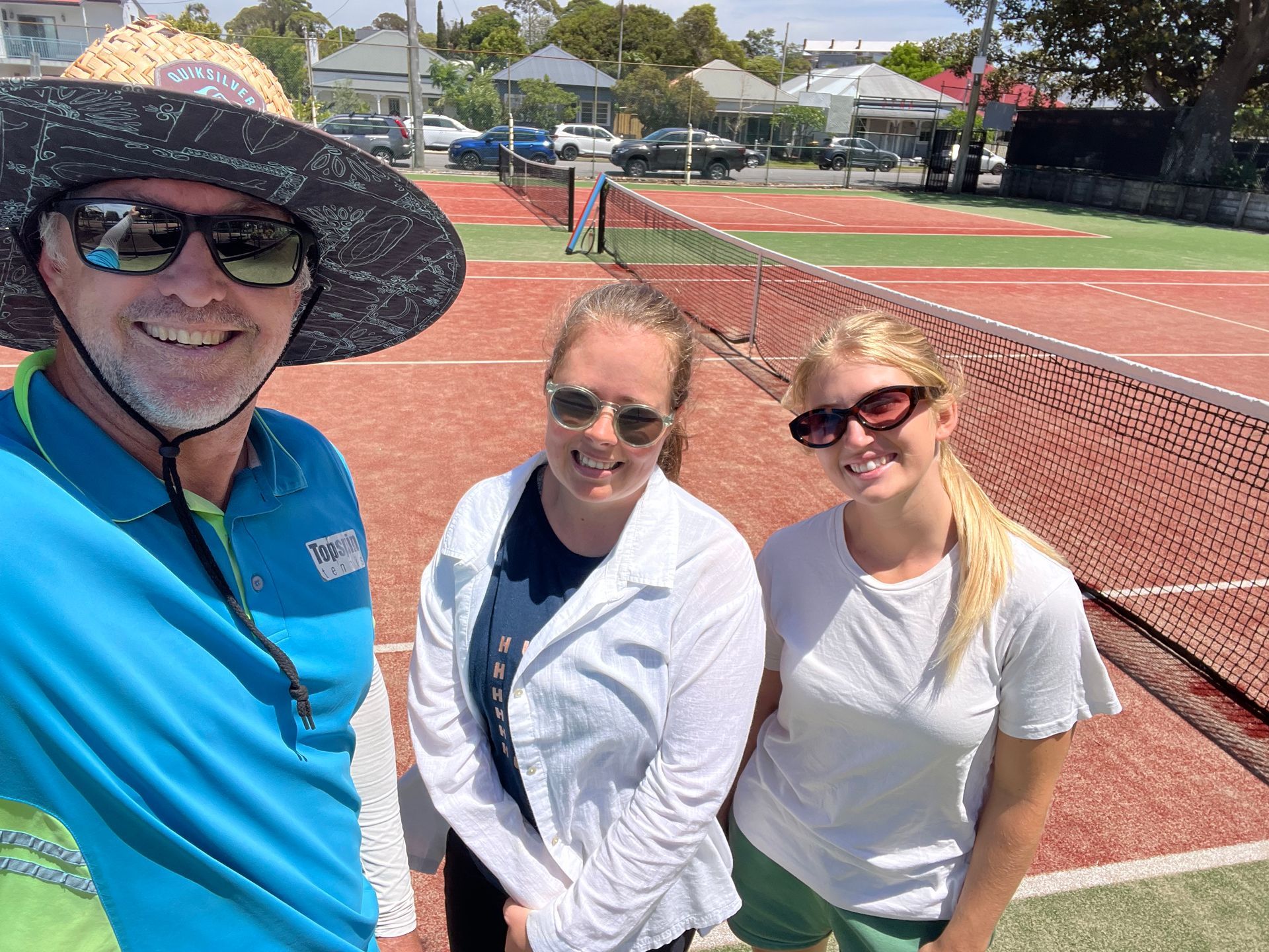 Three People Smiling in Front of a Tennis Net. Man in Hat — Topspin Tennis Newcastle in Lambton, NSW