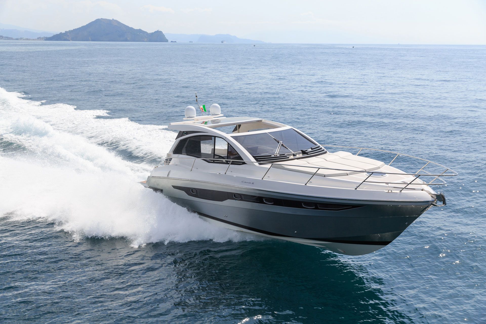 A sleek, silver yacht speeds across the open sea, creating a large white wake against a clear, sunny sky.