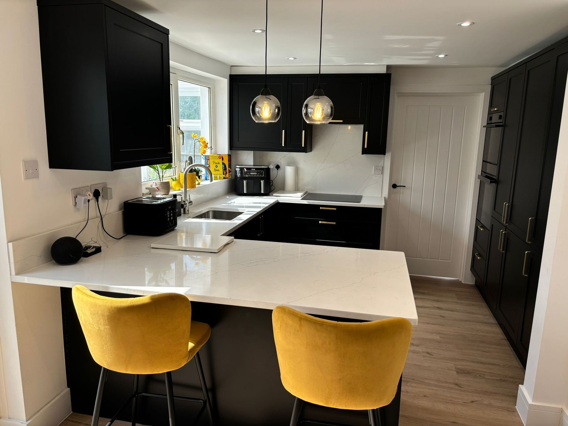 A modern kitchen featuring black cabinetry, white countertops, a breakfast bar with two yellow chairs, and pendant lighting.