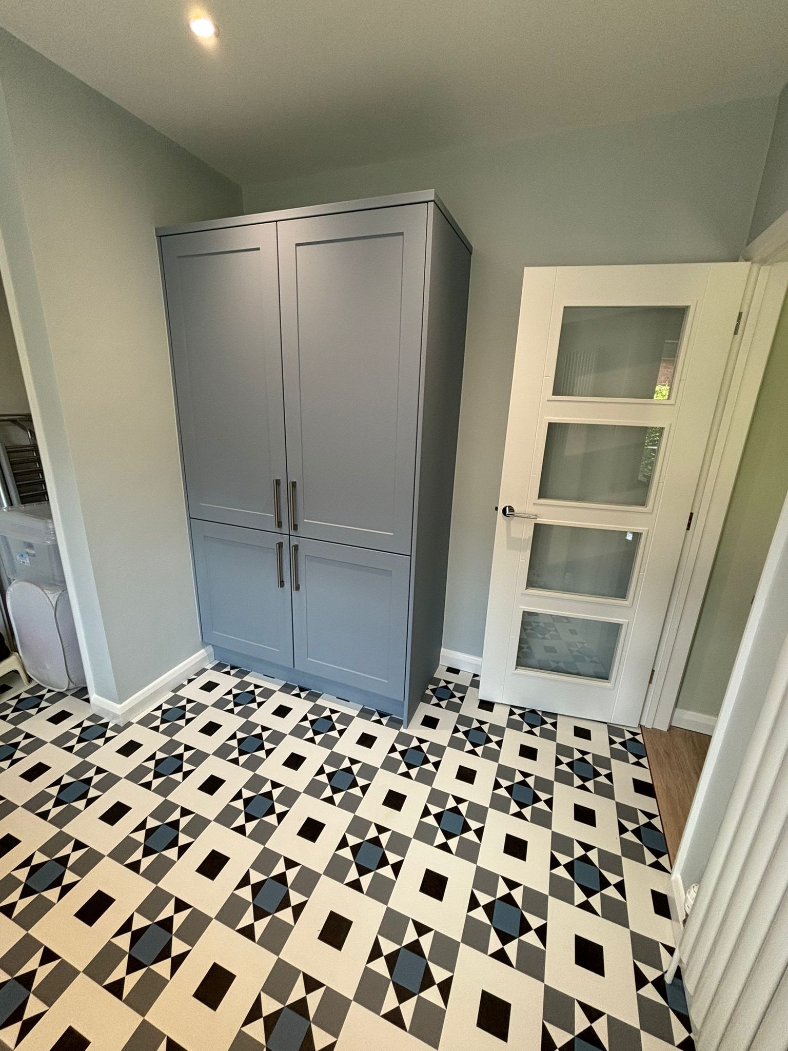 A gray wardrobe stands in a room with patterned geometric floor tiles and a white door with frosted glass panels.