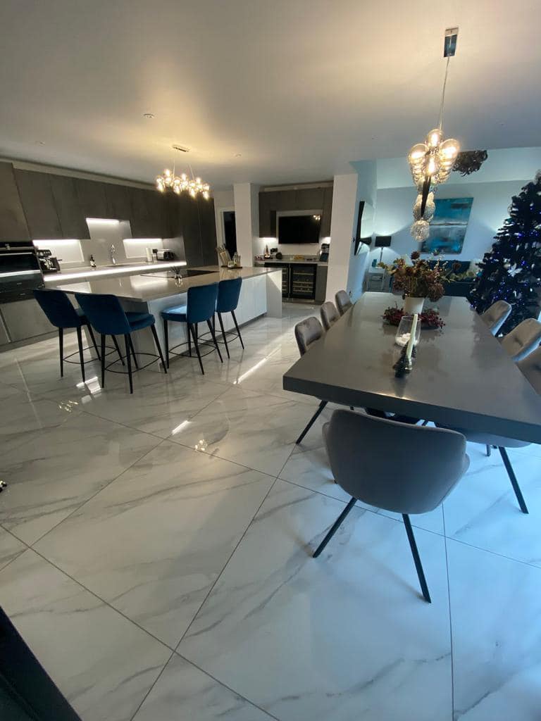 Modern open-plan kitchen and dining area with marble-look floor tiles, grey upholstered chairs, and pendant lighting.
