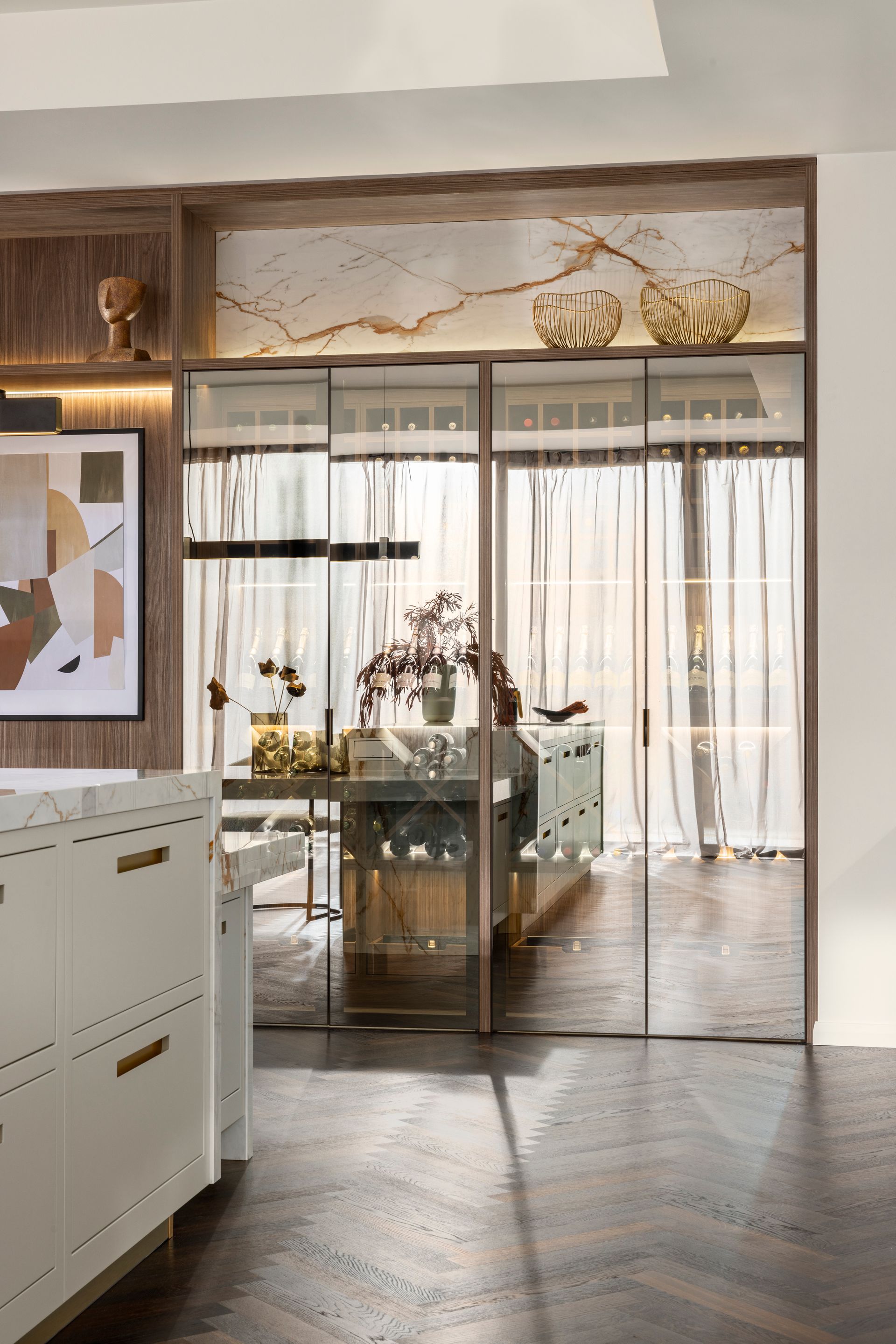 A glass-paneled room divider with gold-trimmed shelving above, set between a white cabinet and a dark herringbone floor.