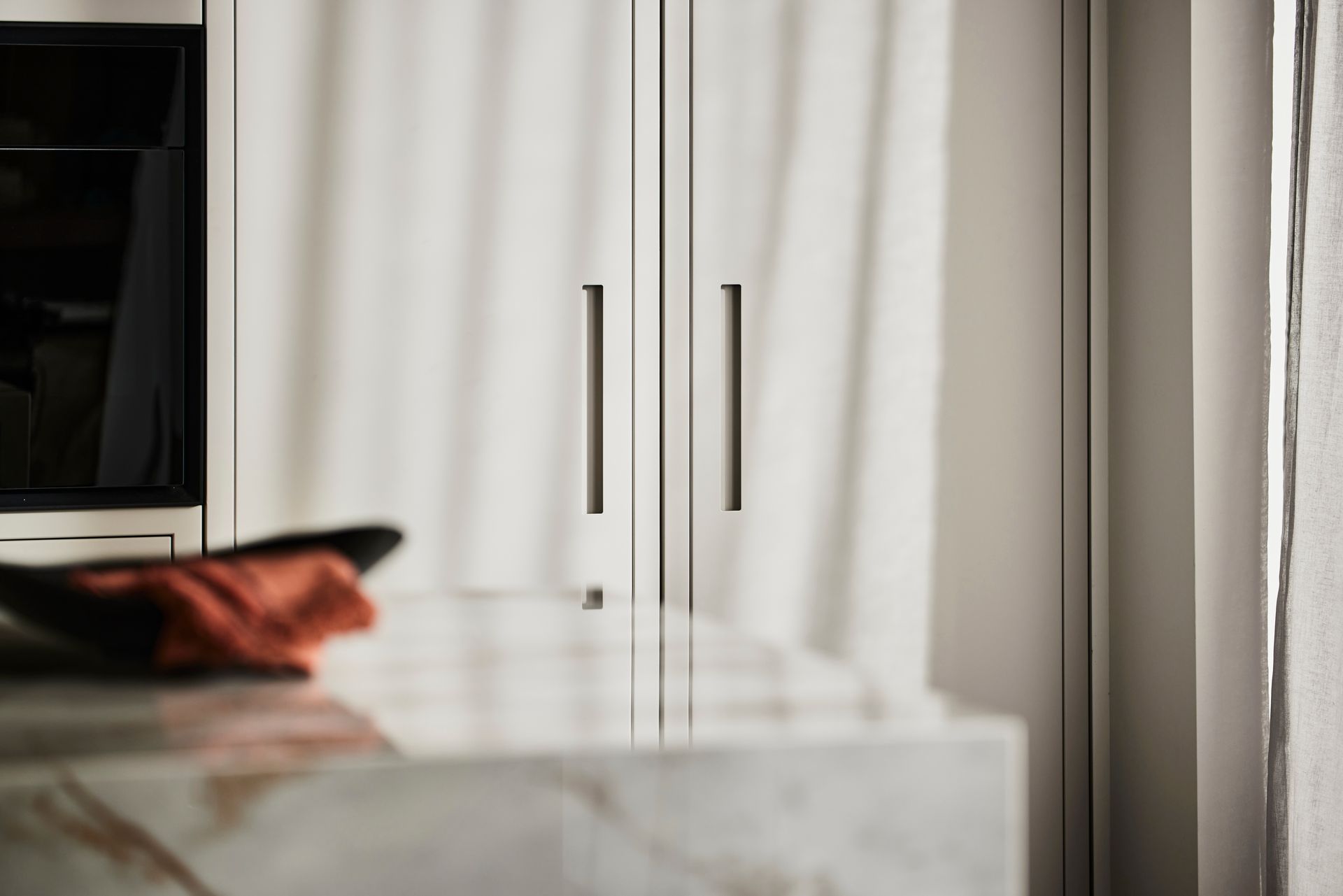 Close-up of white kitchen cabinetry with slim, vertical metal handles, featuring a blurred marble countertop in foreground.