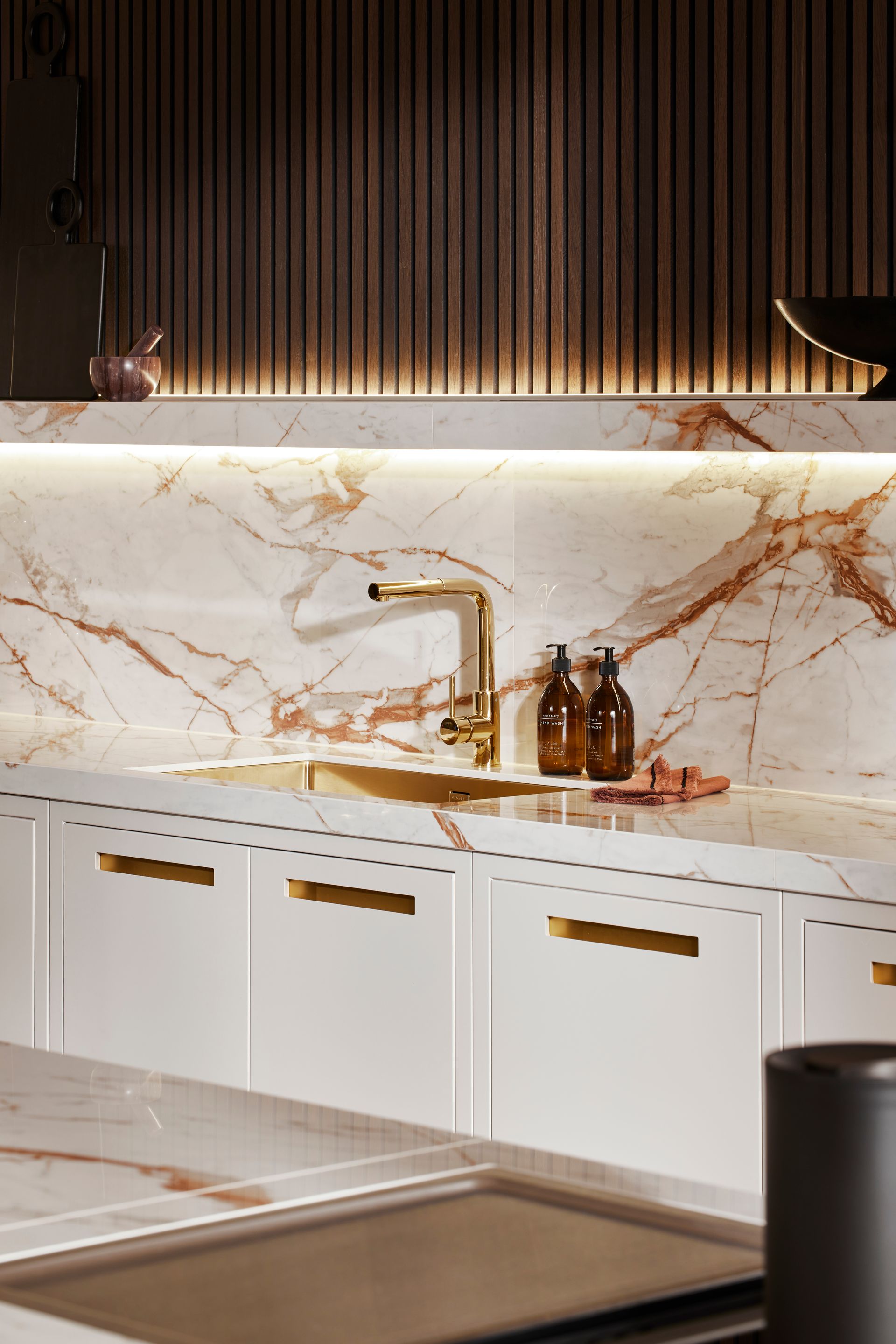 A modern kitchen featuring white cabinets, gold hardware, a marble backsplash with copper veining, and warm underlighting.