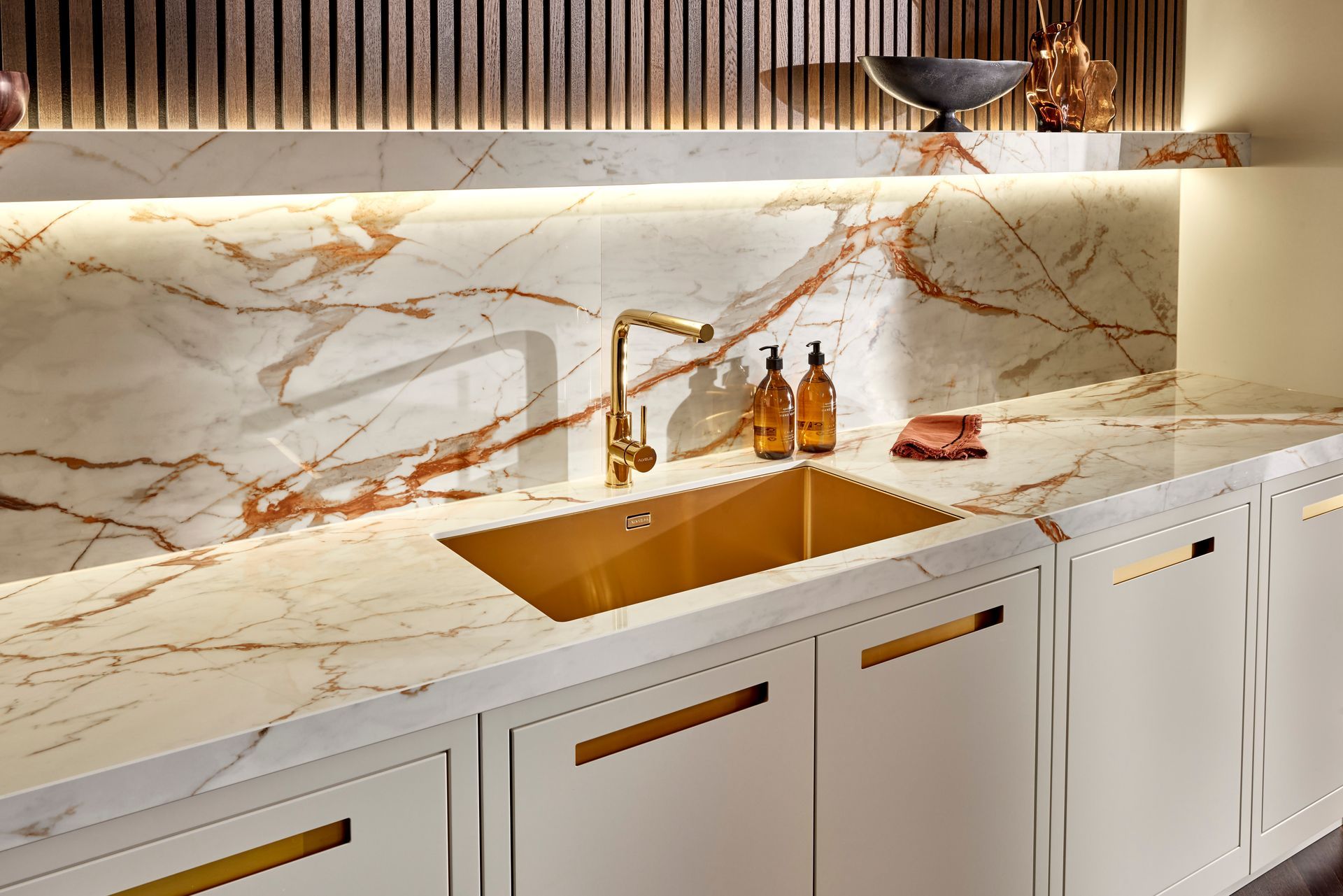A modern kitchen sink area with a gold-colored basin, brass faucet, white marble countertops, and light-colored cabinetry.