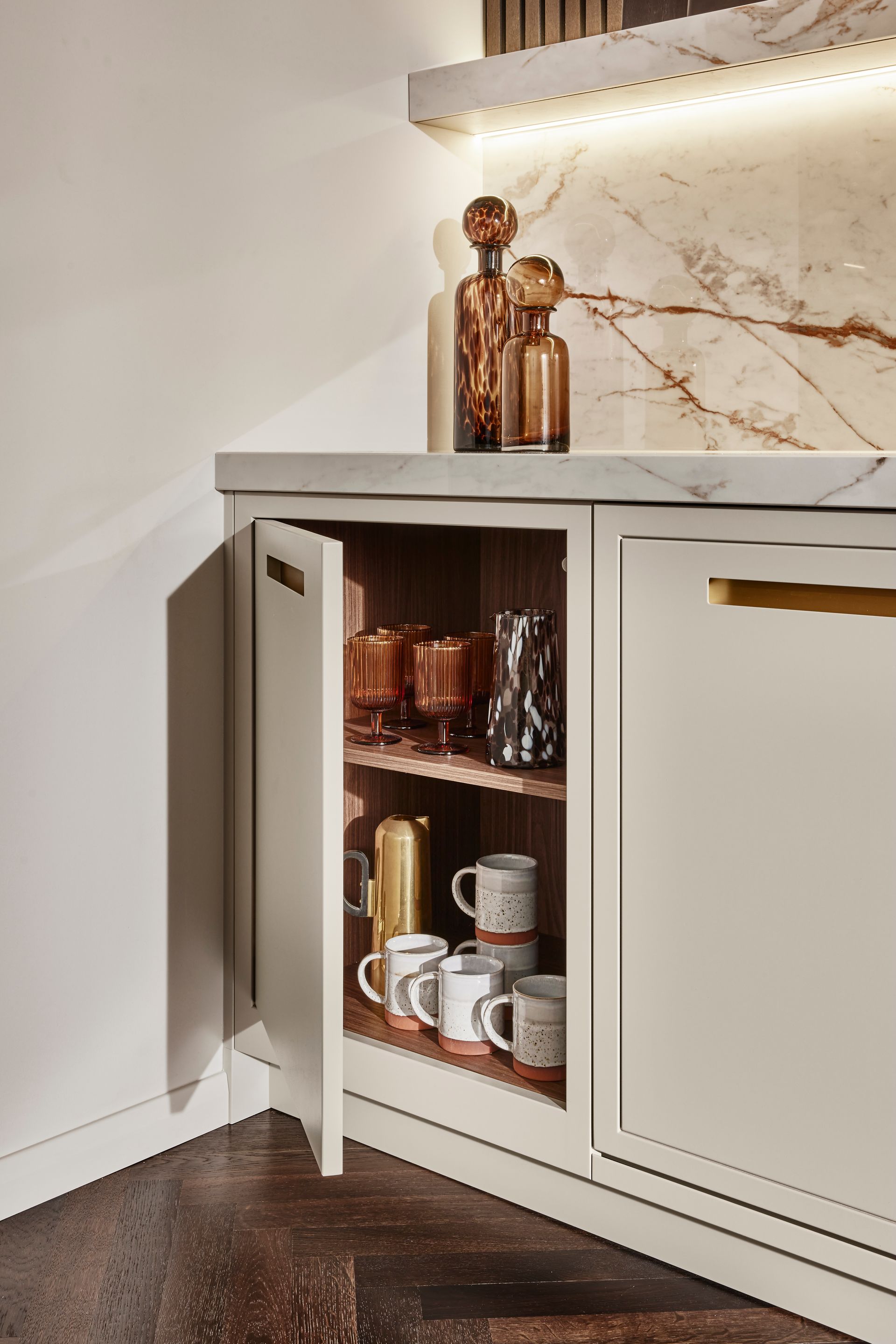 An open cream-colored cabinet door reveals mugs and glass bottles stored on shelves in a kitchen with marble accents.