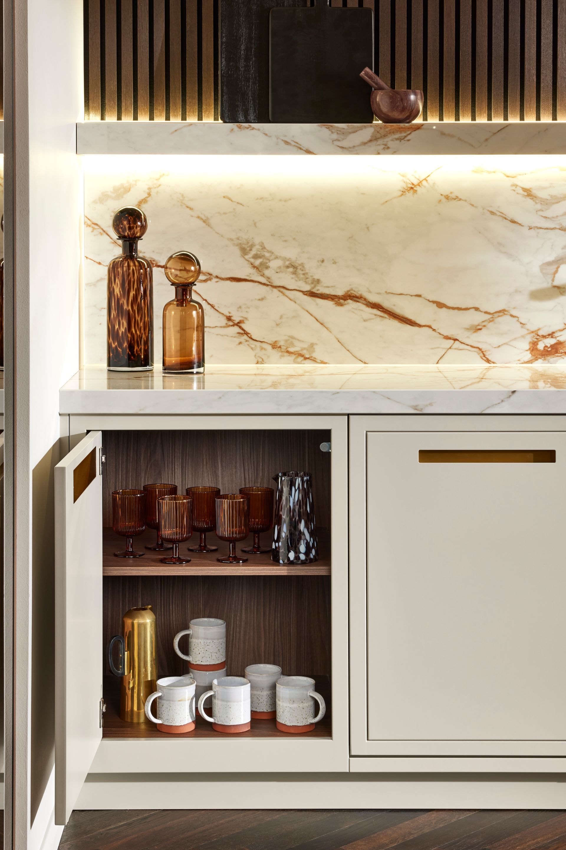 A kitchen cabinet with an open door displays mugs and glassware, under a marble countertop with decorative amber bottles.