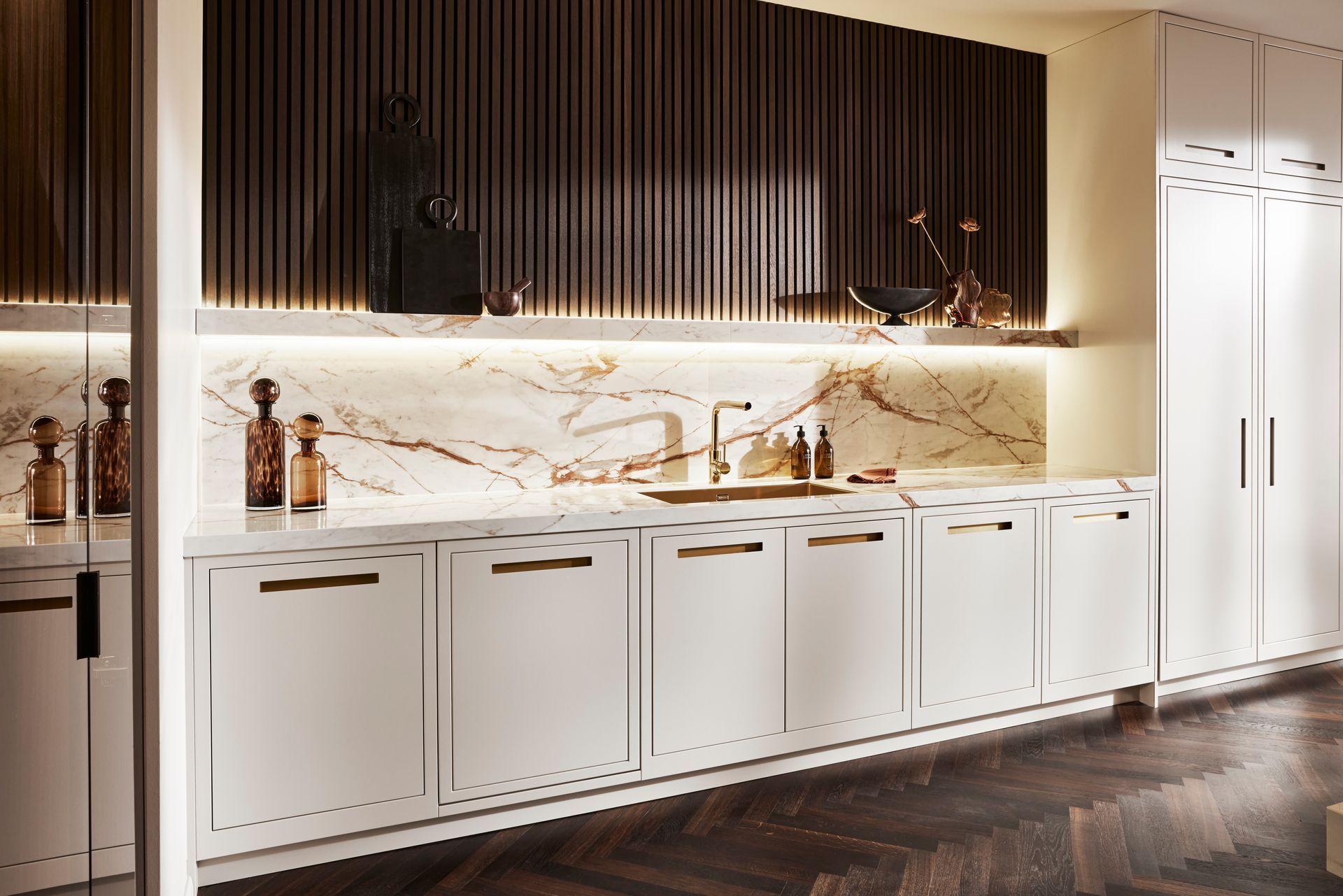 Modern white kitchen with marble backsplash, gold hardware, dark slatted wood walls, and dark herringbone wood floors.