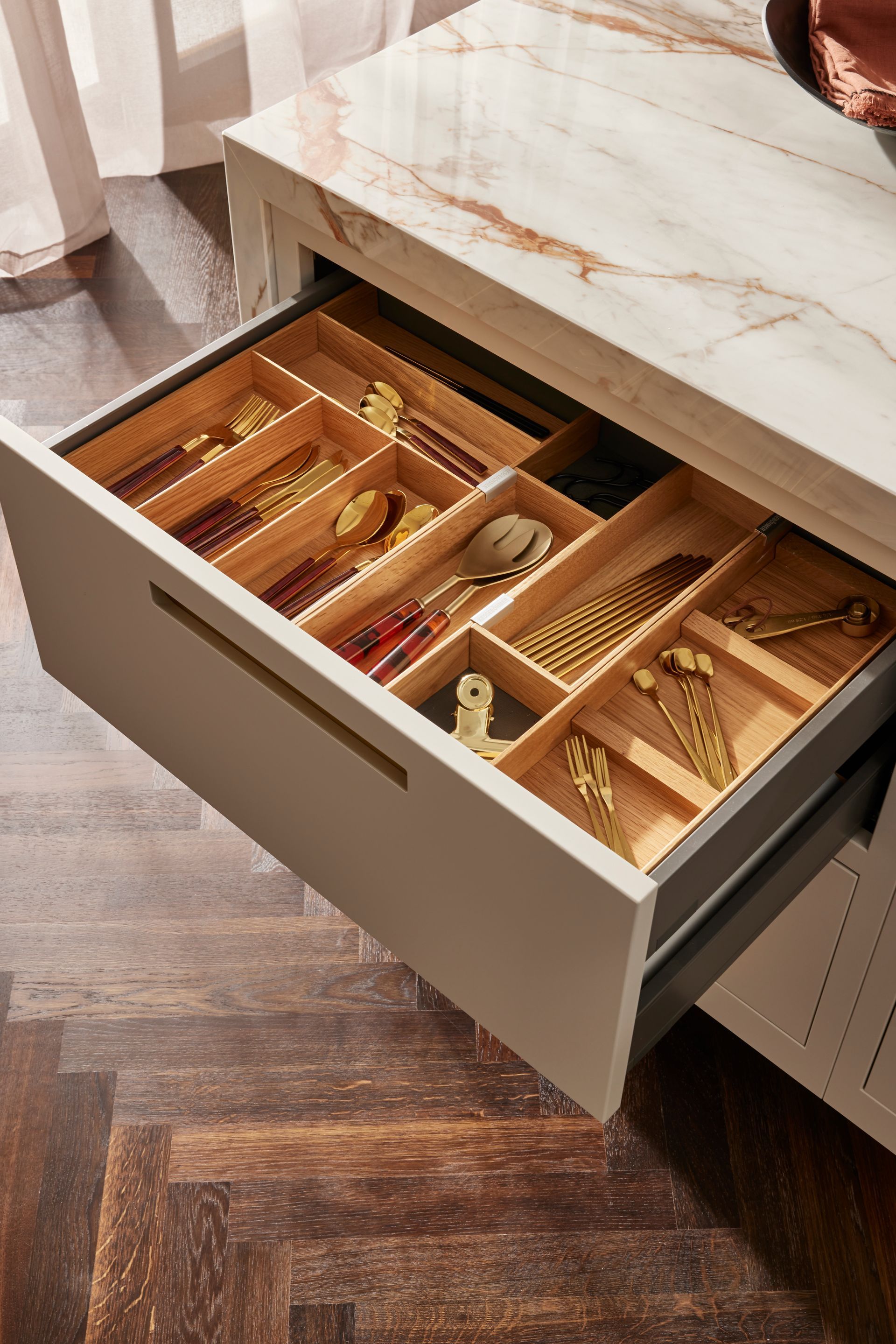 An open drawer with wooden organizers holding gold cutlery, set in a cabinet with a marble countertop on a parquet floor.