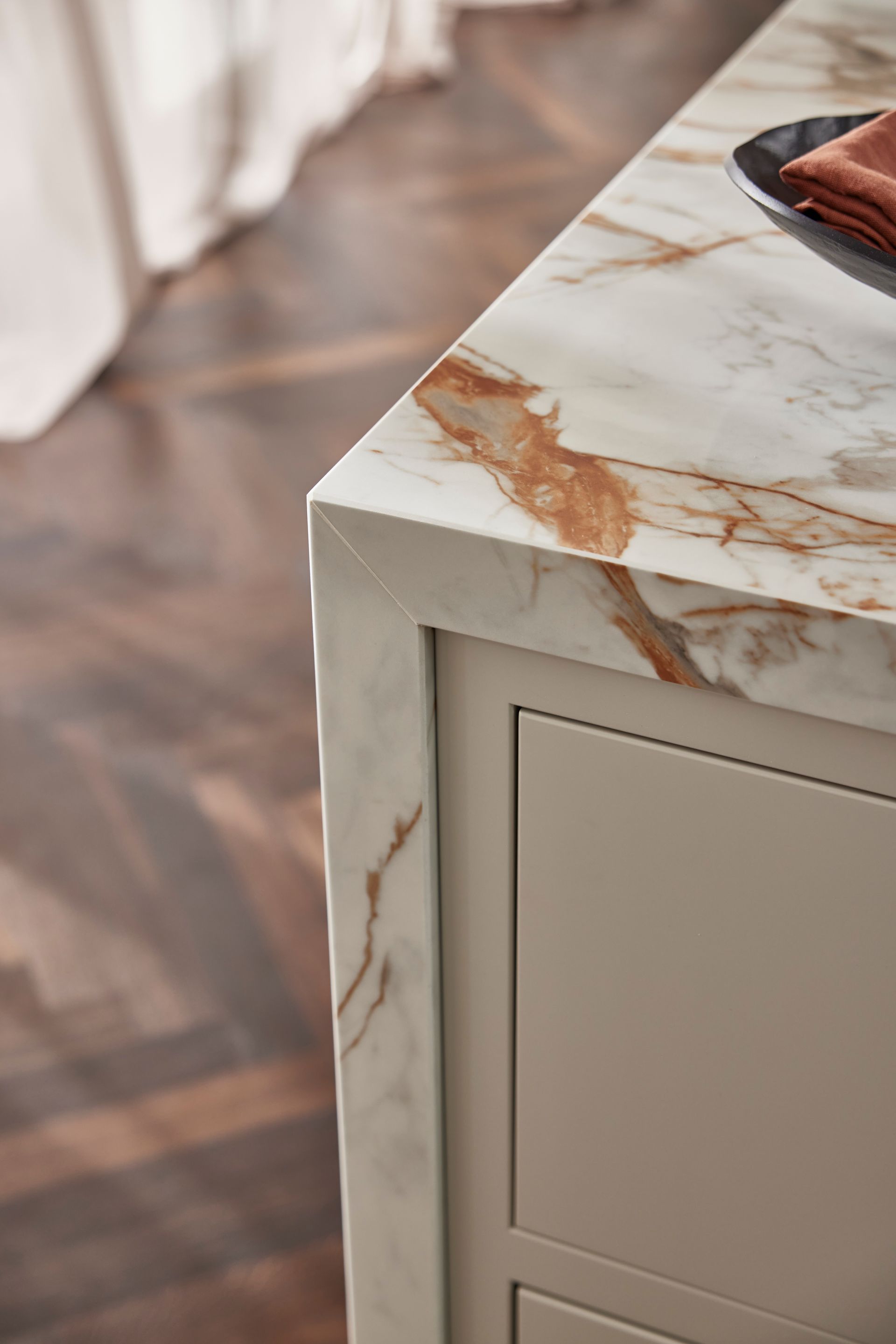 A close-up of a marble countertop with amber veining, featuring a mitered edge corner above light beige cabinetry.