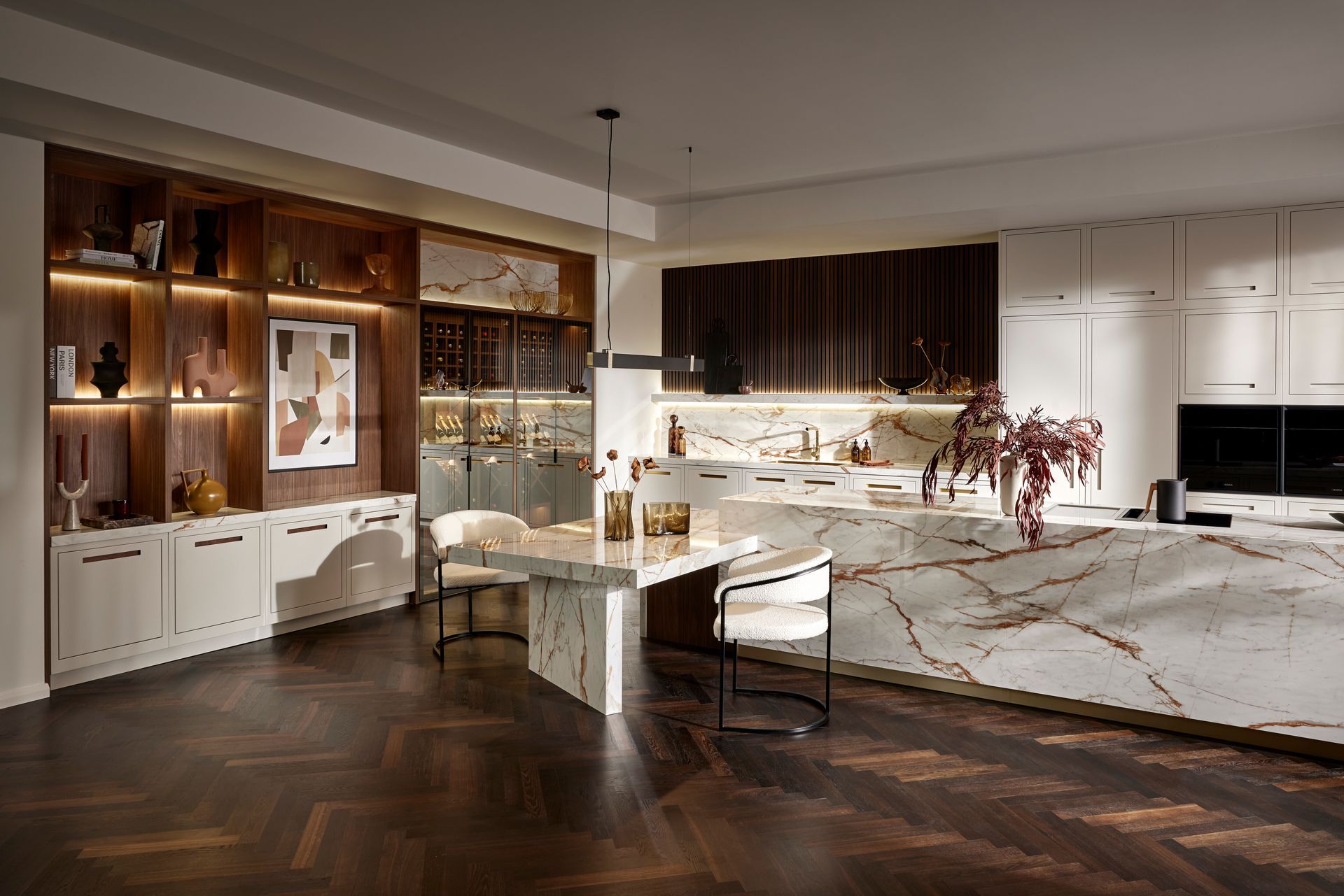 Luxury kitchen with a marble island, dining table, dark wood herringbone floors, and warm ambient lighting.
