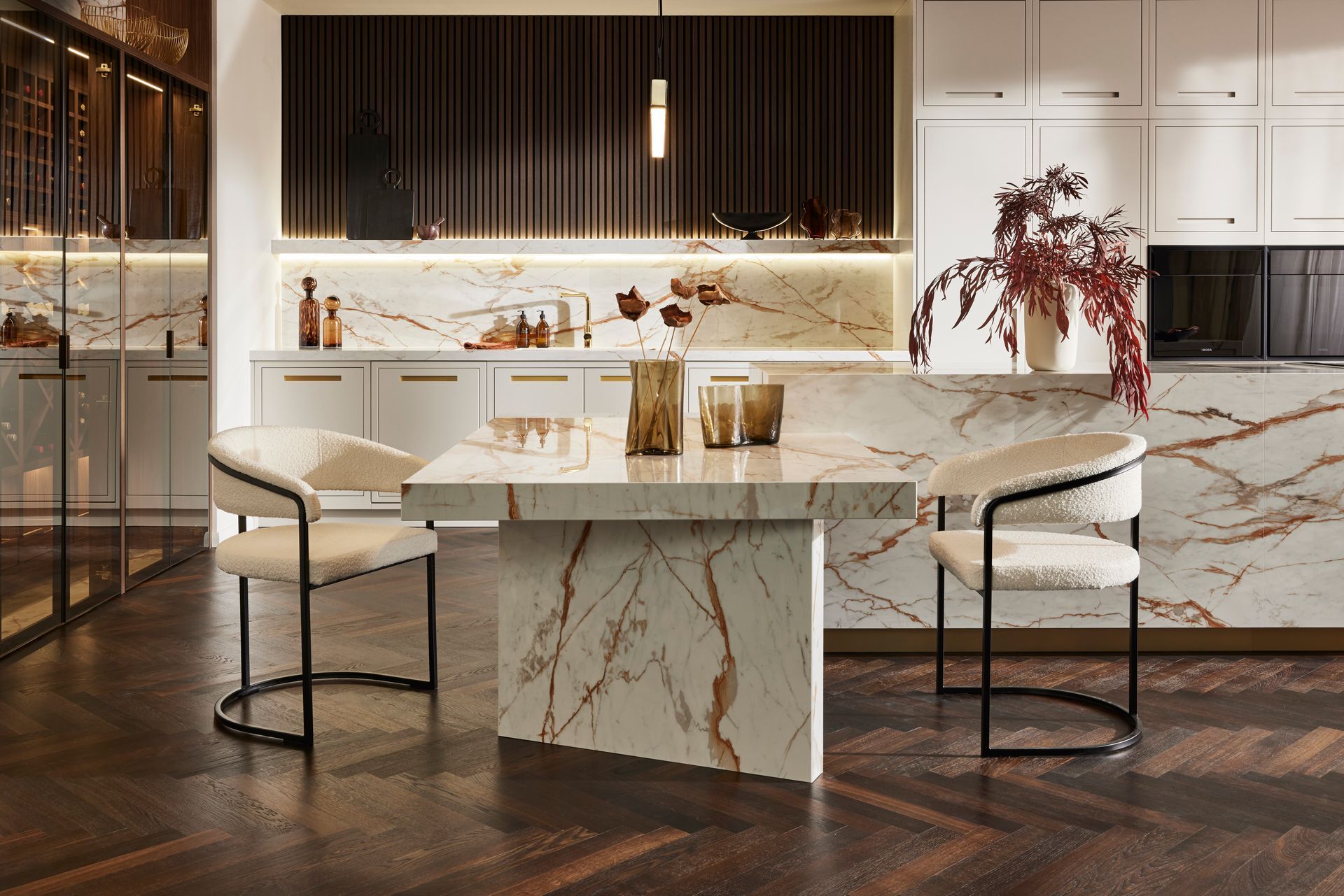 A modern dining area with a white marble table, two upholstered chairs, and a marble kitchen island on dark wood floors.
