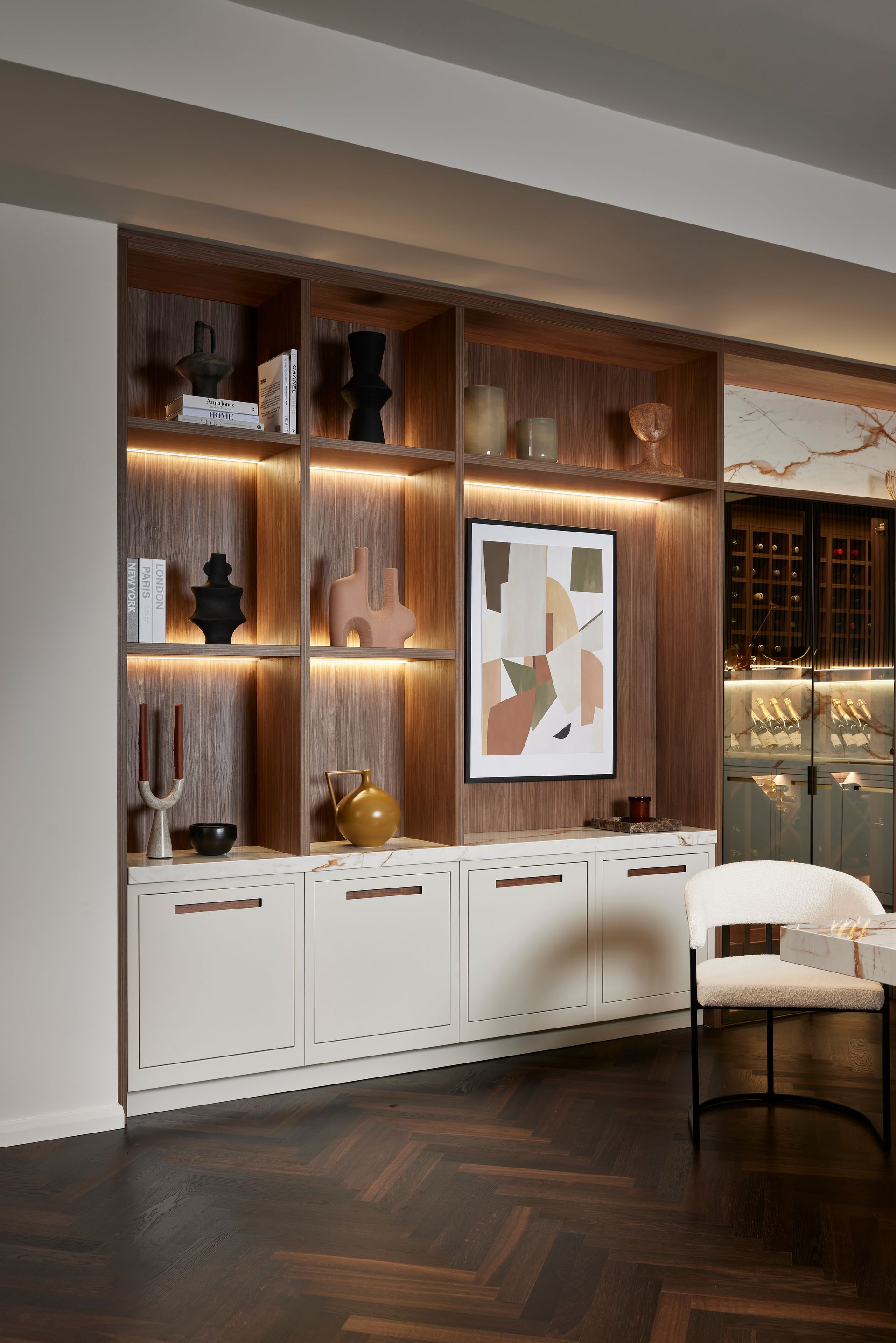 A built-in walnut shelving unit with white cabinets, decorative vases, books, and artwork, illuminated by LED lights.