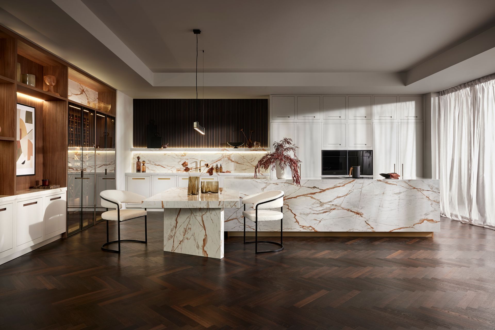 A luxury kitchen with a white marble island and table, wood-paneled accents, and a dark herringbone floor.