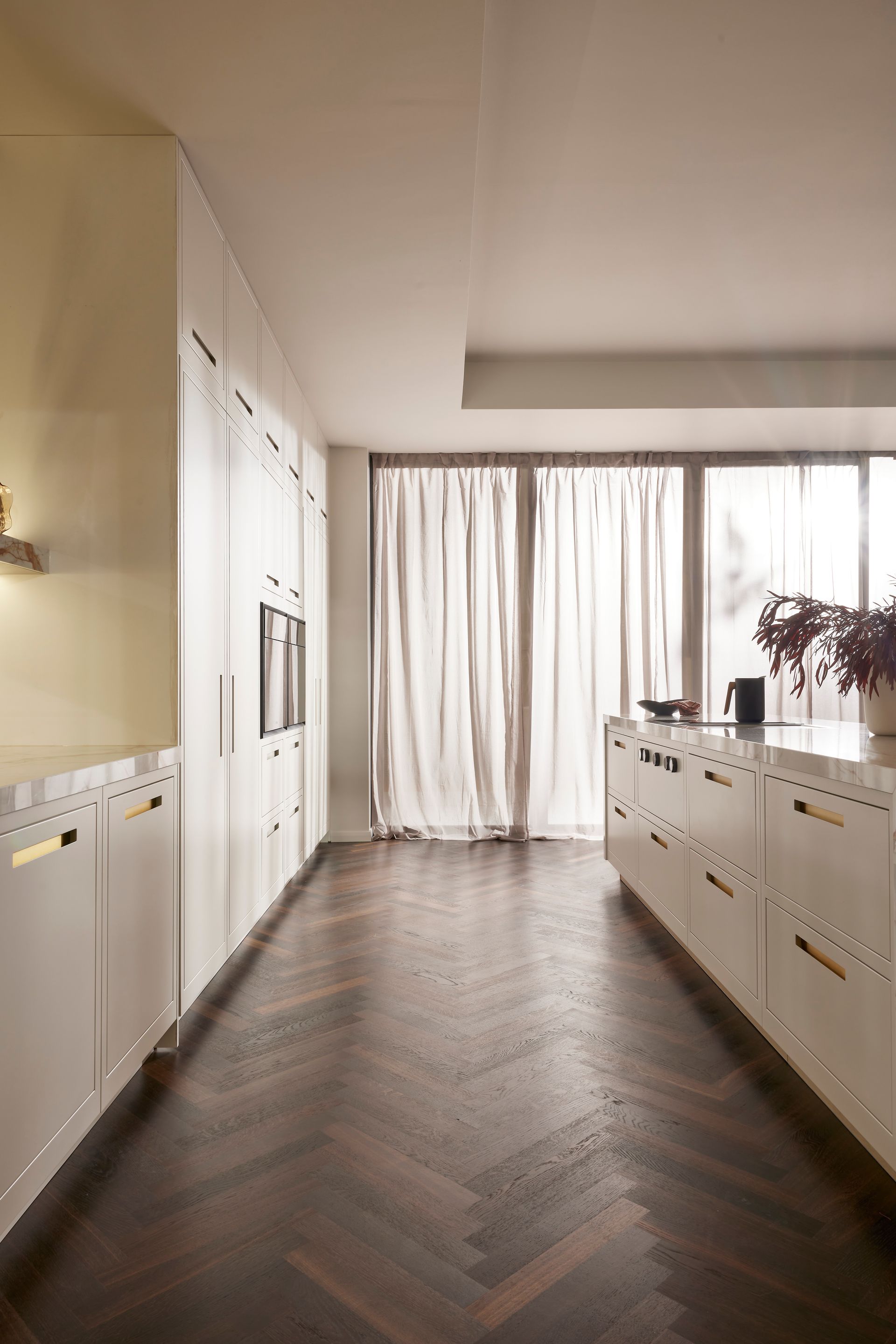 A modern kitchen with white cabinets, gold handles, and a dark herringbone wood floor, facing a bright curtained window.