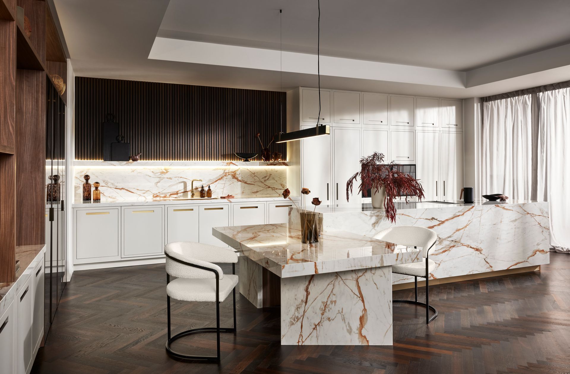 Modern kitchen with dark wood cabinets, white marble island, dining table, and white chairs on a dark herringbone floor.