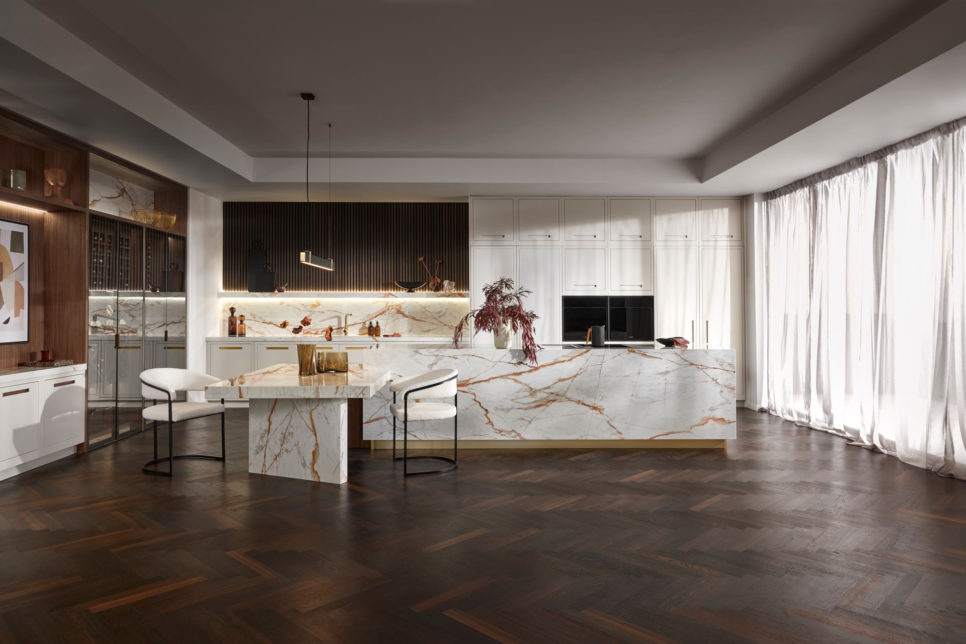 A modern kitchen with a white marble island, dark wood herringbone floors, and warm ambient lighting.