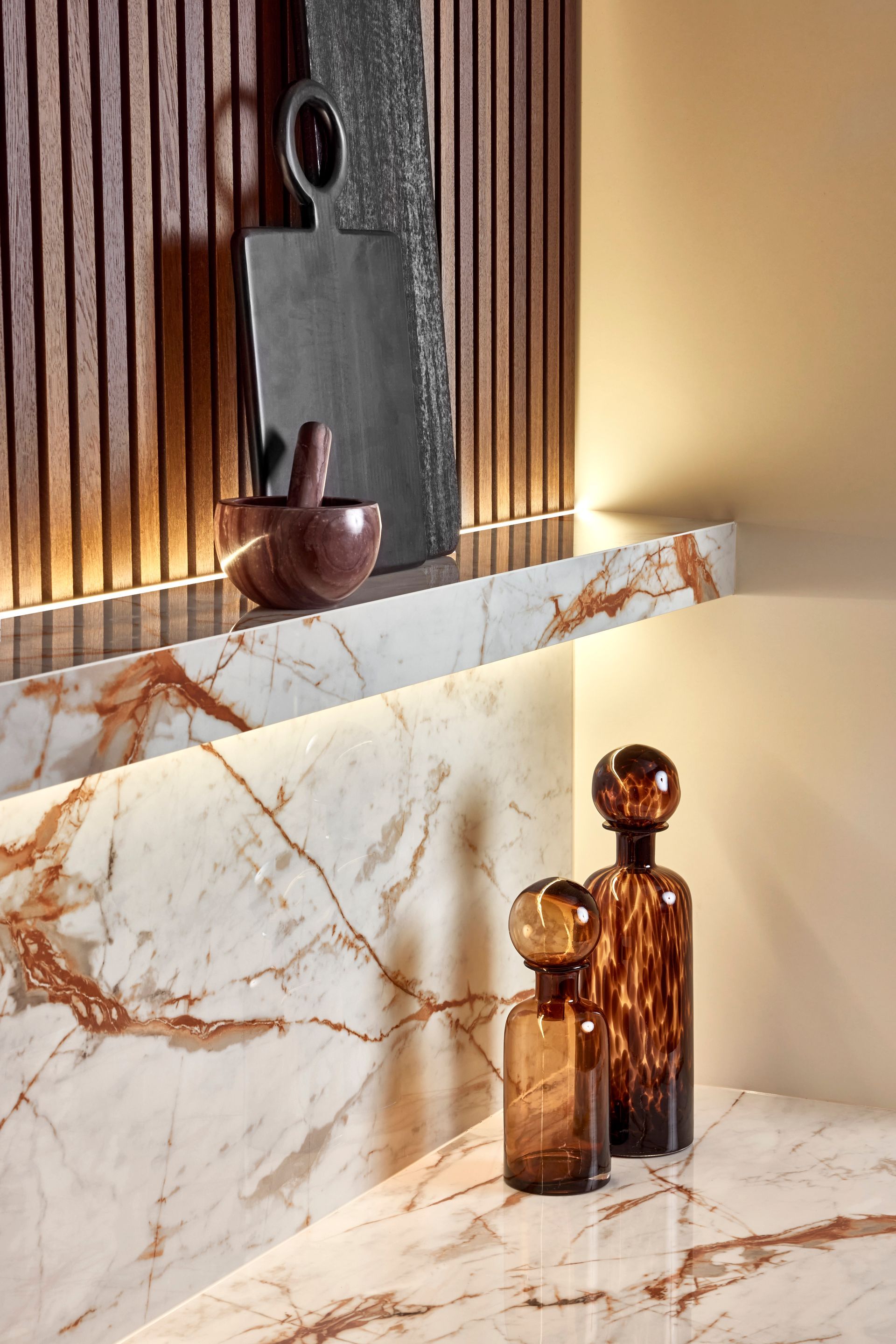 A marble shelf and countertop feature amber-tinted glass bottles, a dark stone board, and a small, spherical bowl.