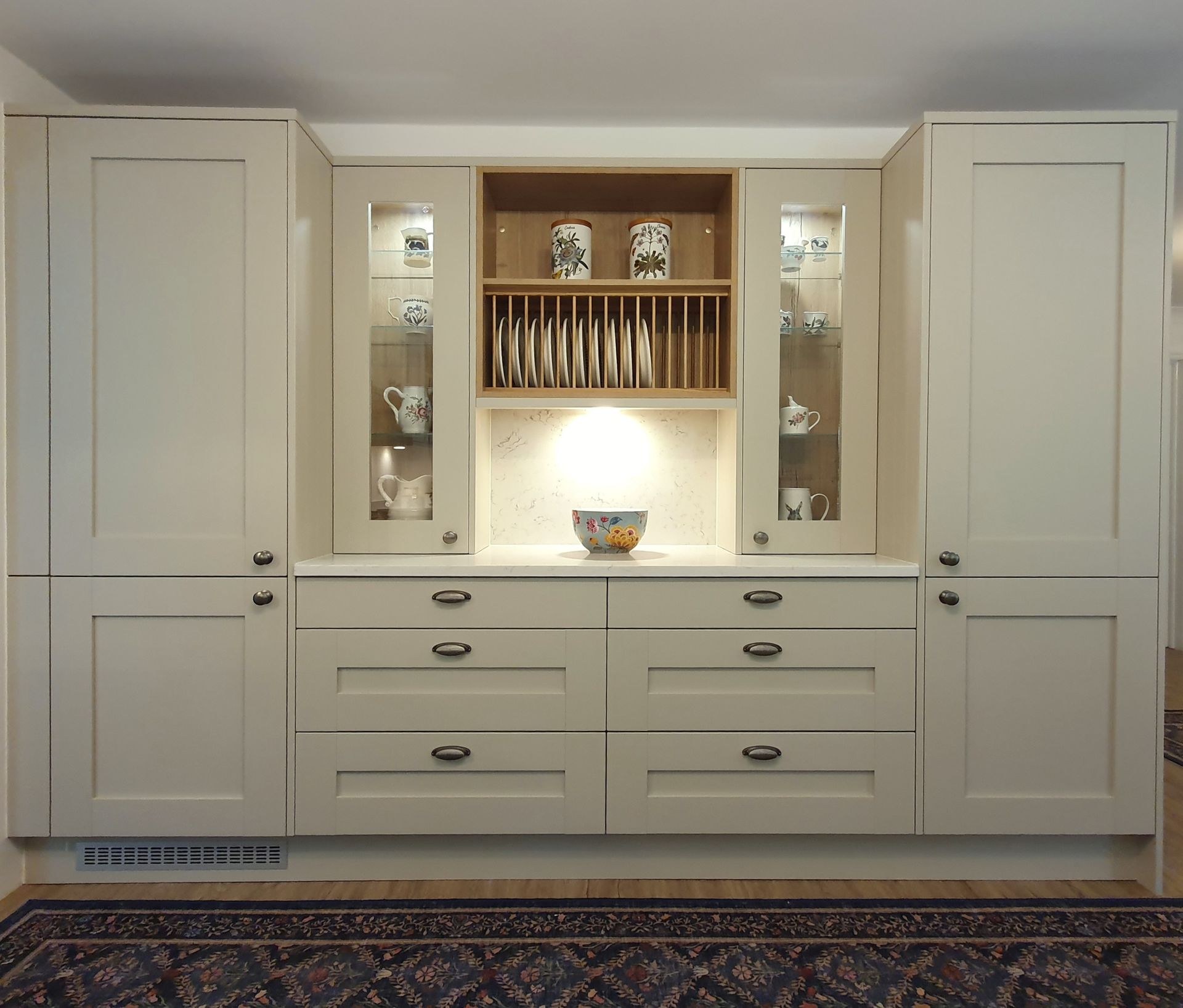 A built-in cream-colored kitchen hutch featuring side cabinets, glass-fronted shelves, a plate rack, and base drawers.
