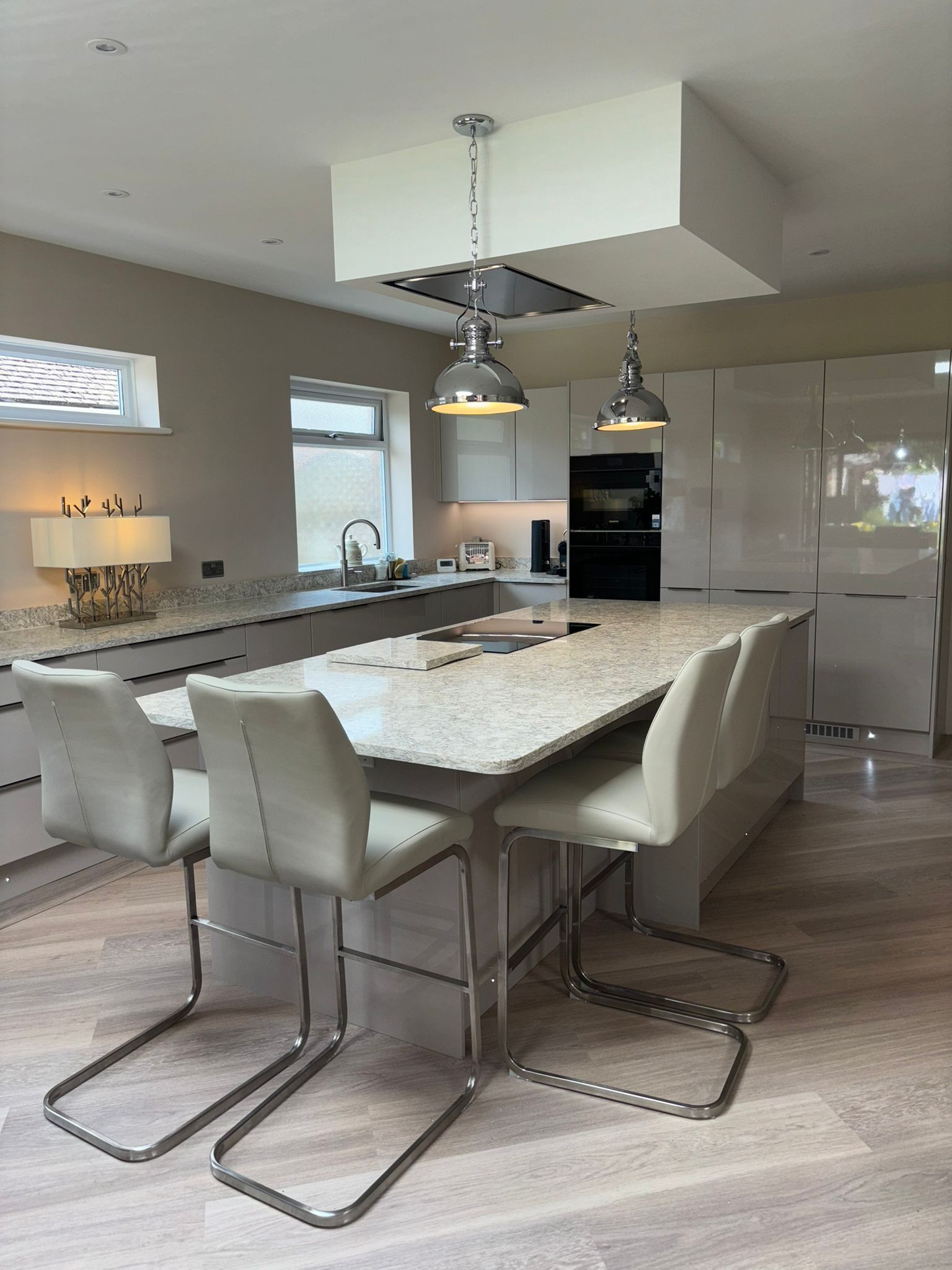 Modern kitchen featuring a marble-topped island with three beige bar stools under pendant lights and light wood floors.