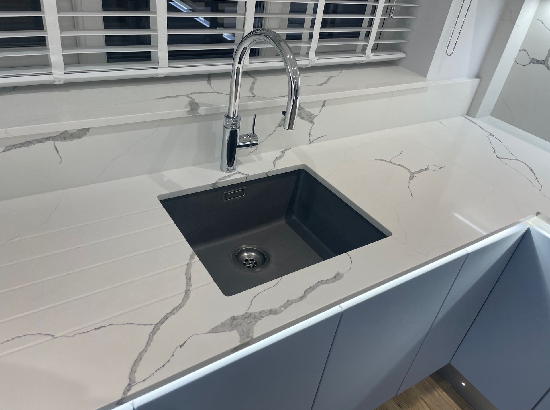 A chrome faucet over a dark square sink set in a white, gray-veined marble countertop with blue cabinets below.