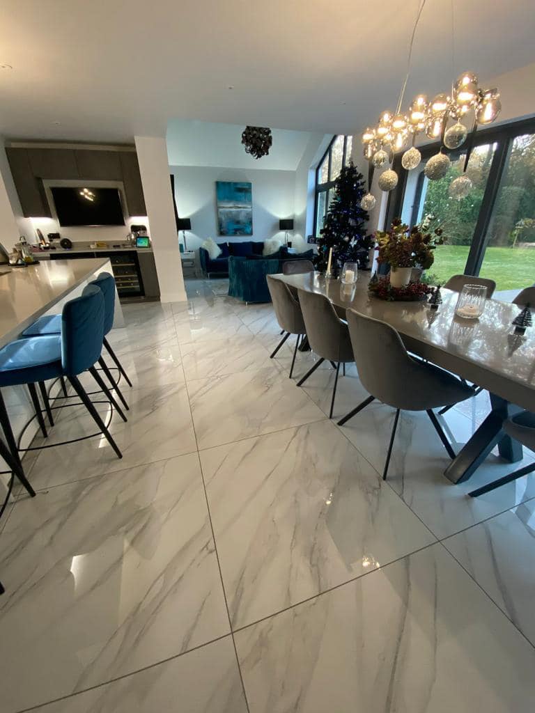 A modern dining and kitchen area with marble-look tile floors, a long grey table, blue bar stools, and a glass chandelier.