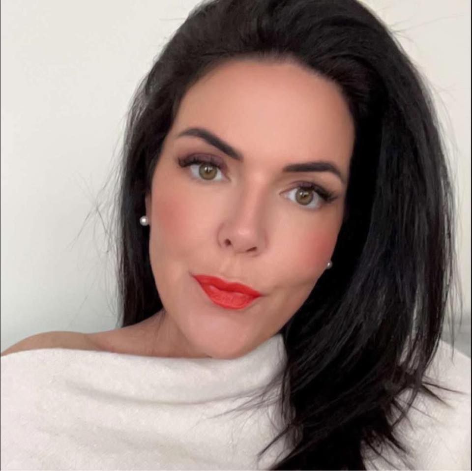 A person with long dark hair and red lipstick poses for a headshot while wearing a white off-the-shoulder top.