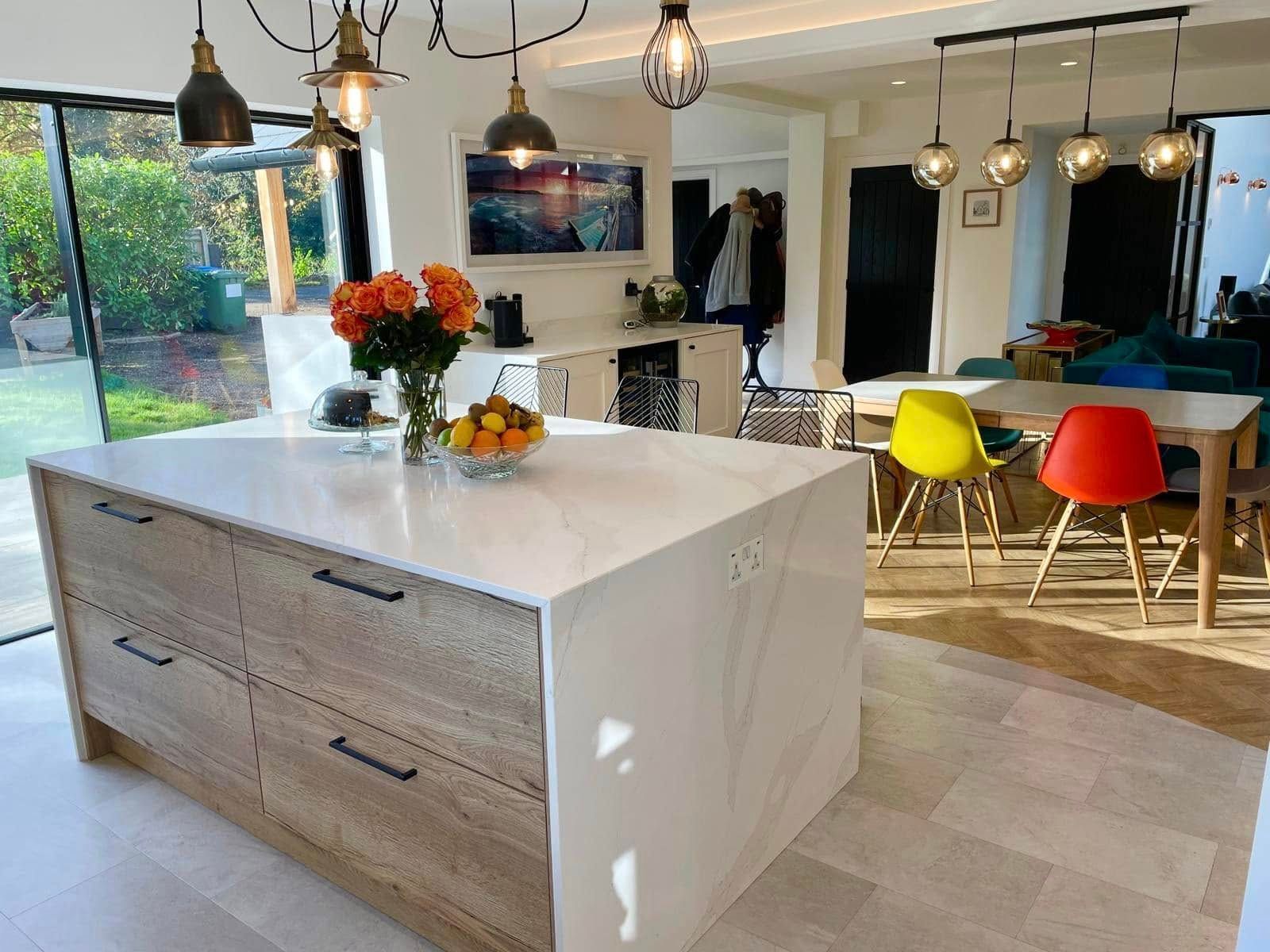 A modern kitchen with a light-wood island, white stone countertop, orange flowers, and bright red and yellow chairs.