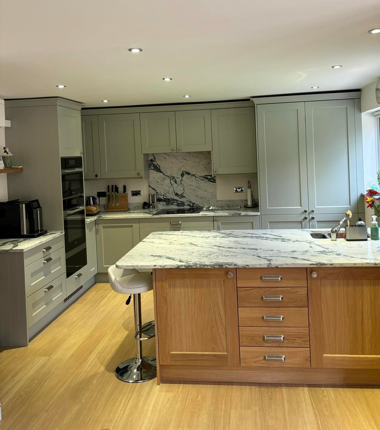 A kitchen with sage green cabinets, a wood-toned island with marble countertops, and light wood flooring.