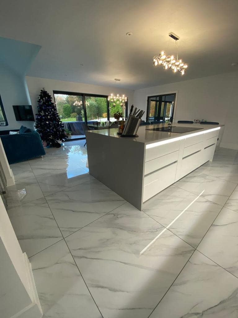 A modern, open-plan kitchen featuring a large gray island, white marble-patterned flooring, and a Christmas tree nearby.
