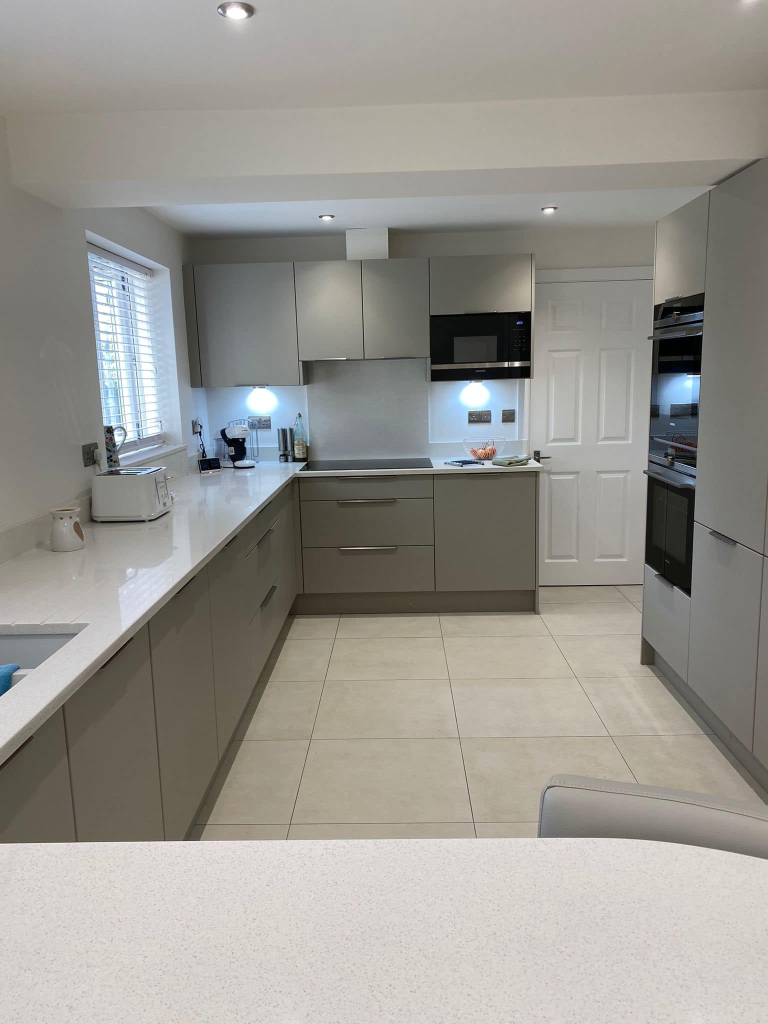 Modern L-shaped kitchen featuring grey cabinets, light countertops, integrated appliances, and a tiled floor.