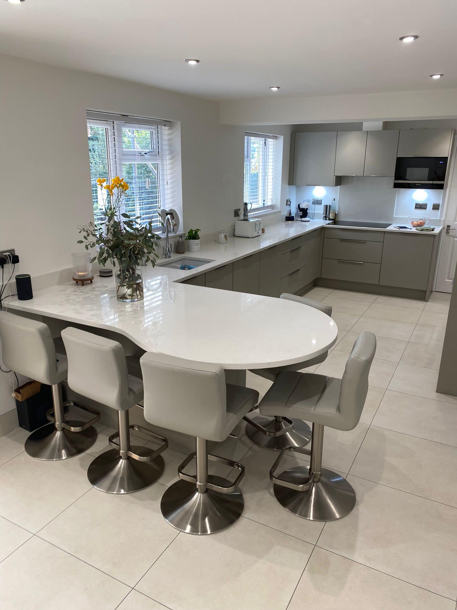 Modern kitchen with a white curved breakfast bar, four grey stools, light tiled flooring, and taupe cabinets.