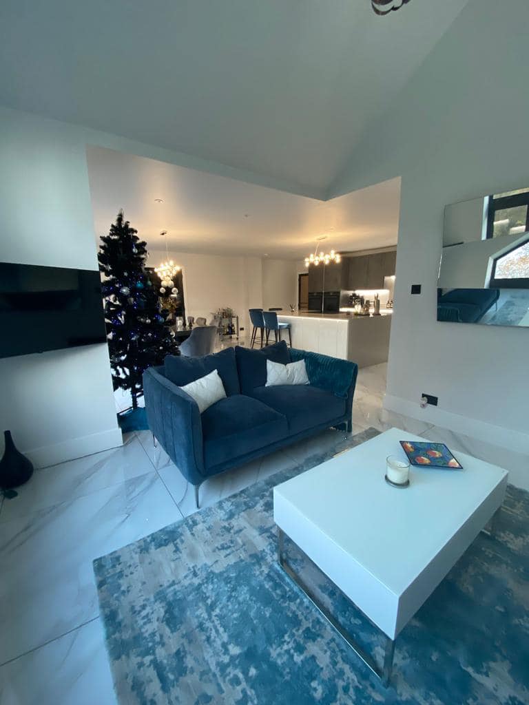 A modern living room with a blue velvet sofa, white coffee table, and area rug, leading to an open-plan kitchen area.