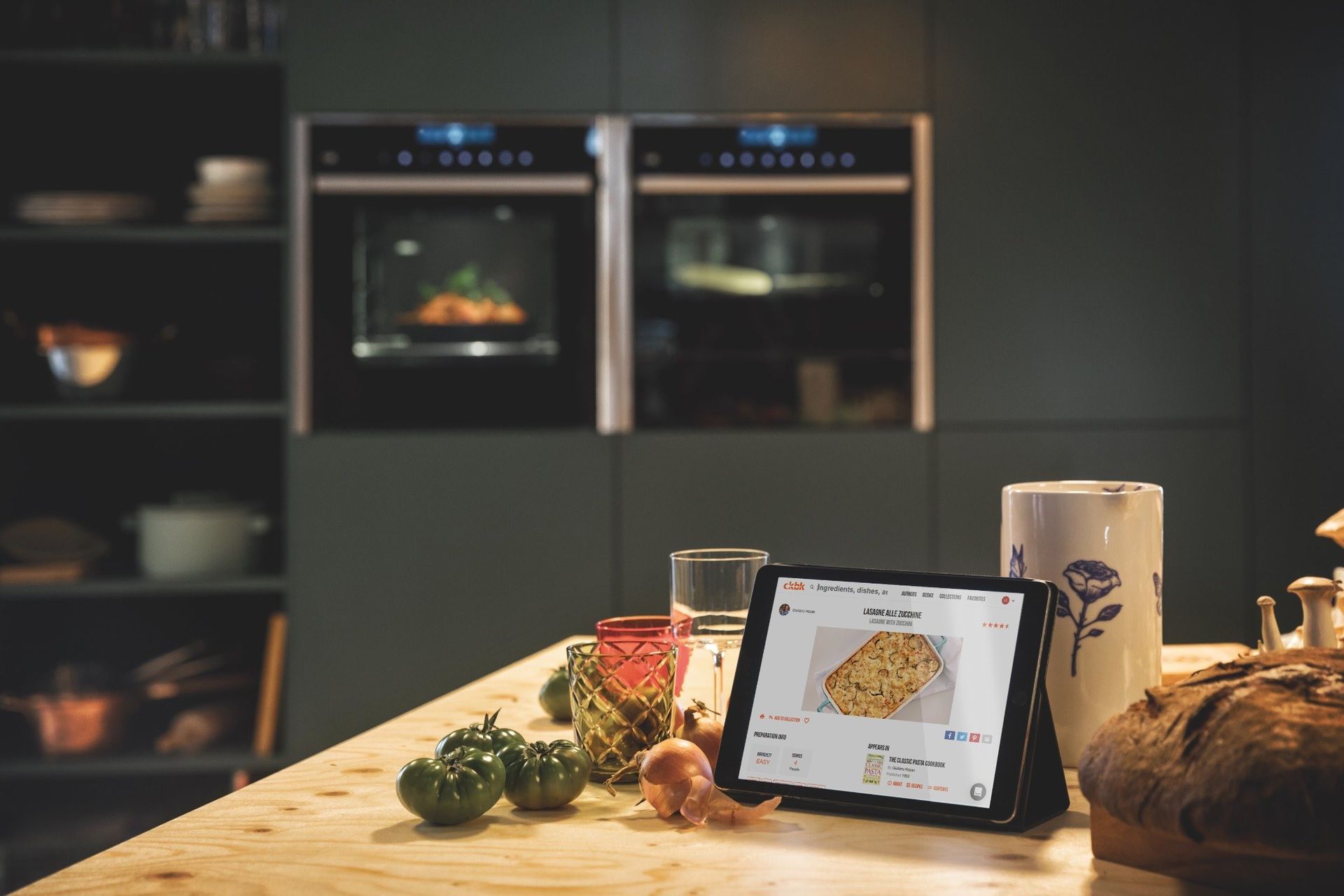 A tablet displaying a cooking app sits on a wooden kitchen counter in front of two ovens, with tomatoes and bread nearby.
