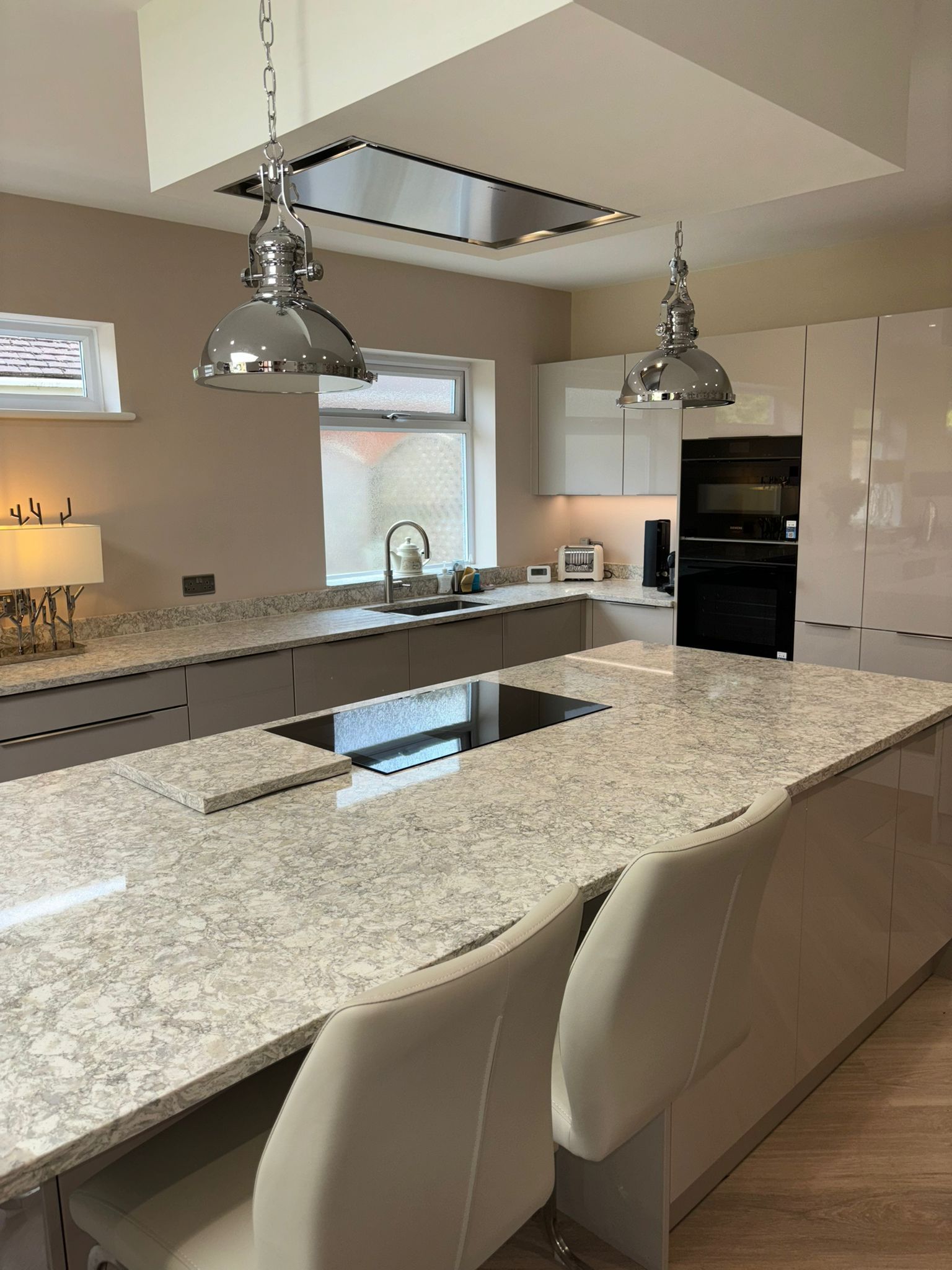 A modern kitchen with a large quartz island, light-colored cabinetry, two chrome pendant lights, and a built-in oven.