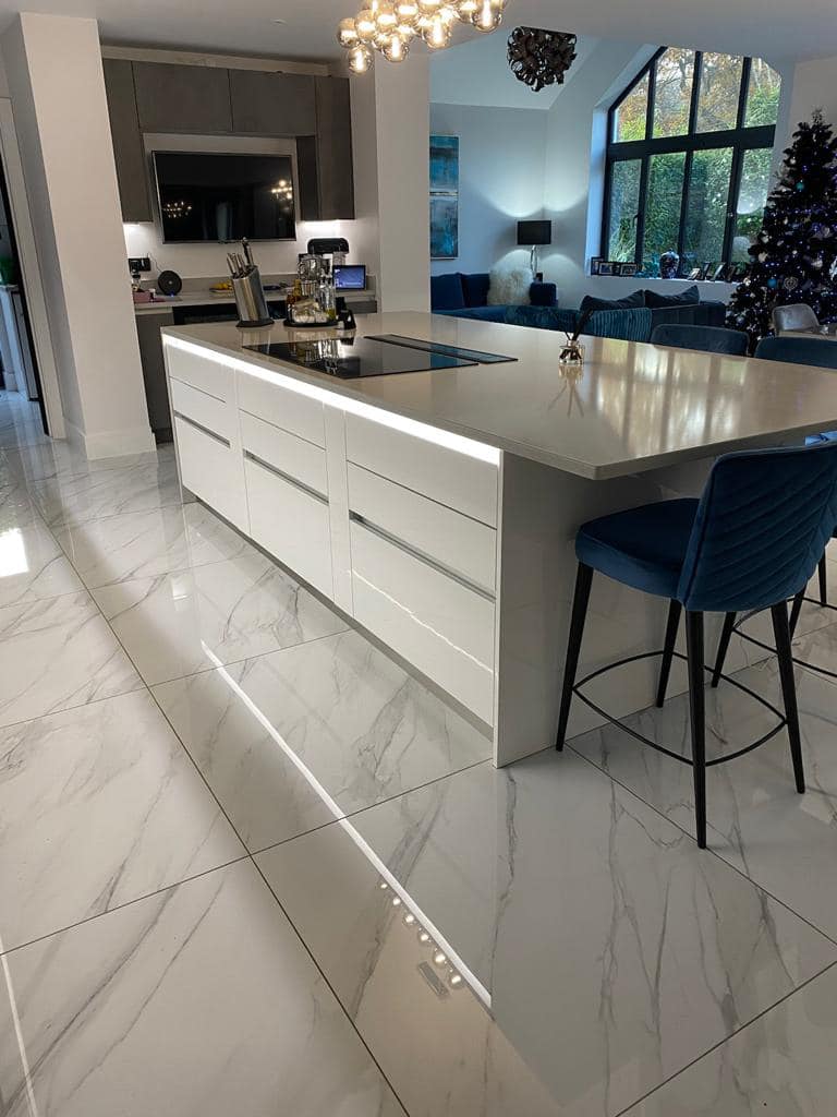 A bright modern kitchen with a large white island, blue bar stool, marble-style flooring, and a Christmas tree.
