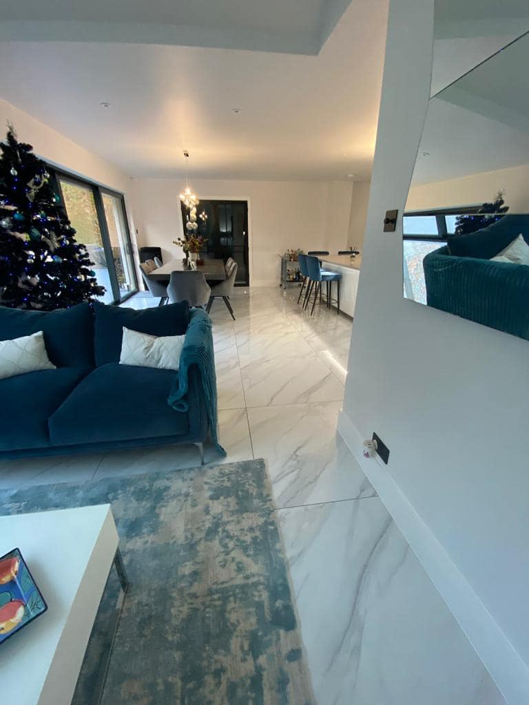 A bright, open-plan living room with white marble floors, a teal sofa, a Christmas tree, and a dining area in the back.