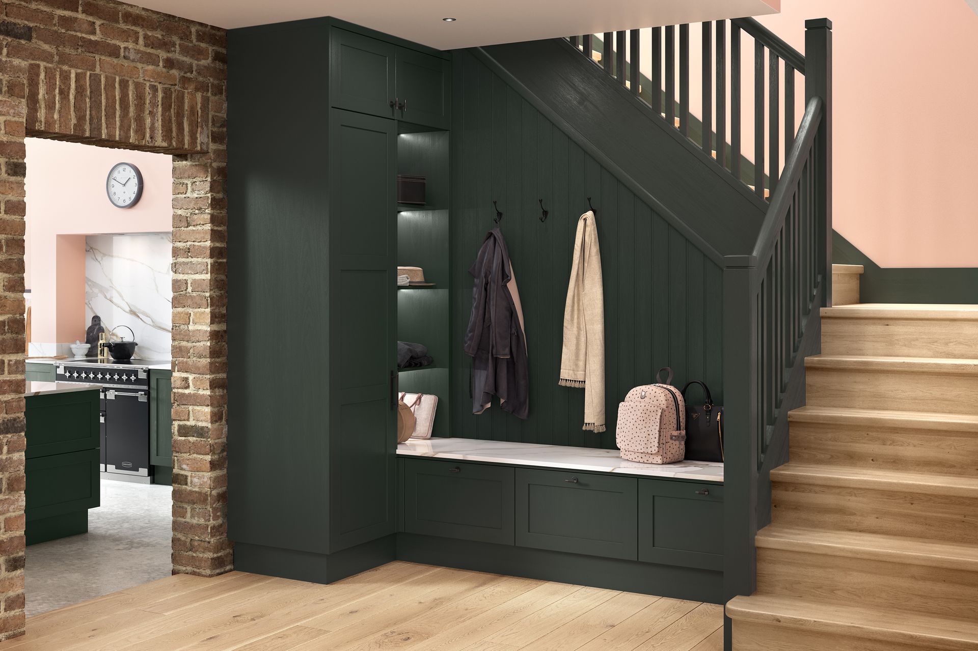 Dark green under-stair mudroom bench with hooks, coats, and bags beside a staircase and brick archway