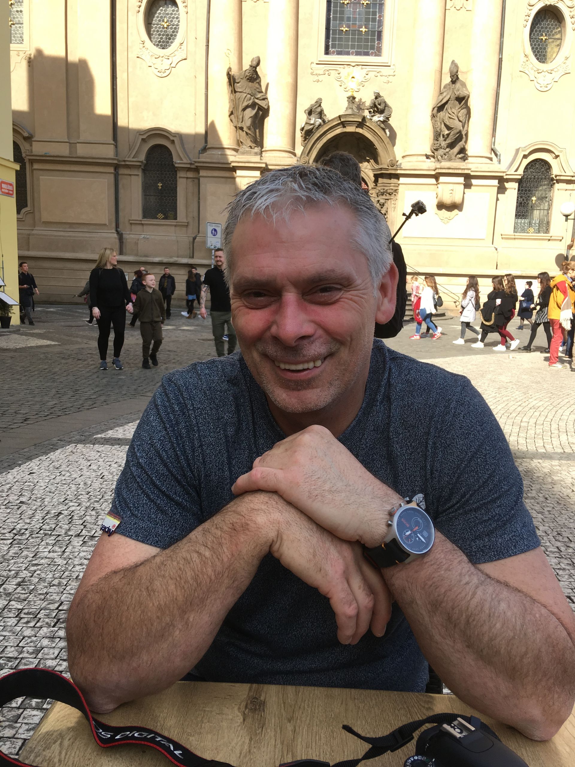 A smiling man with graying hair sits at a table in a cobblestone square in front of a historic building with statues.